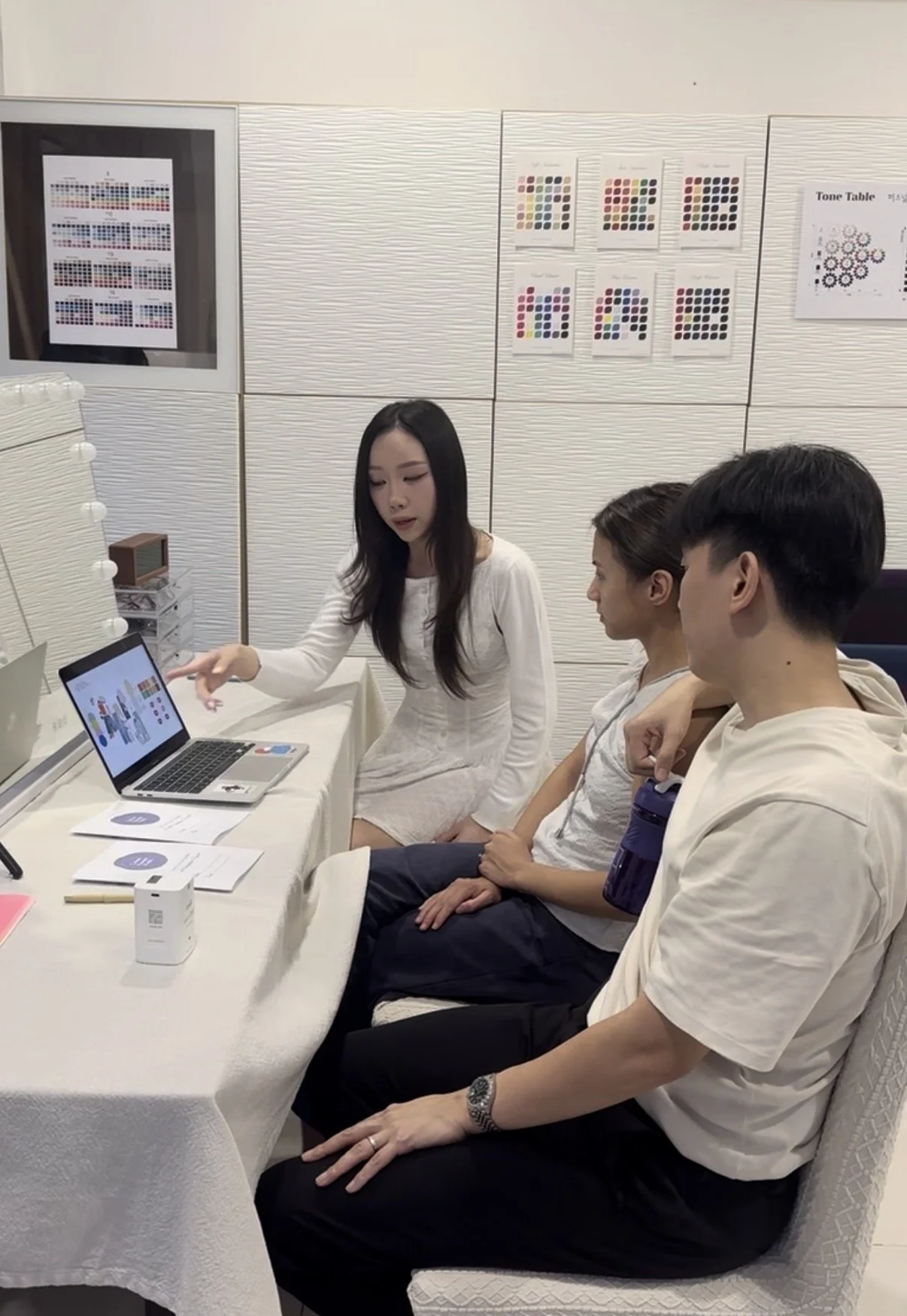 Three people sitting at a table, looking at a laptop screen with a woman pointing at it, in a learning or presentation setting.