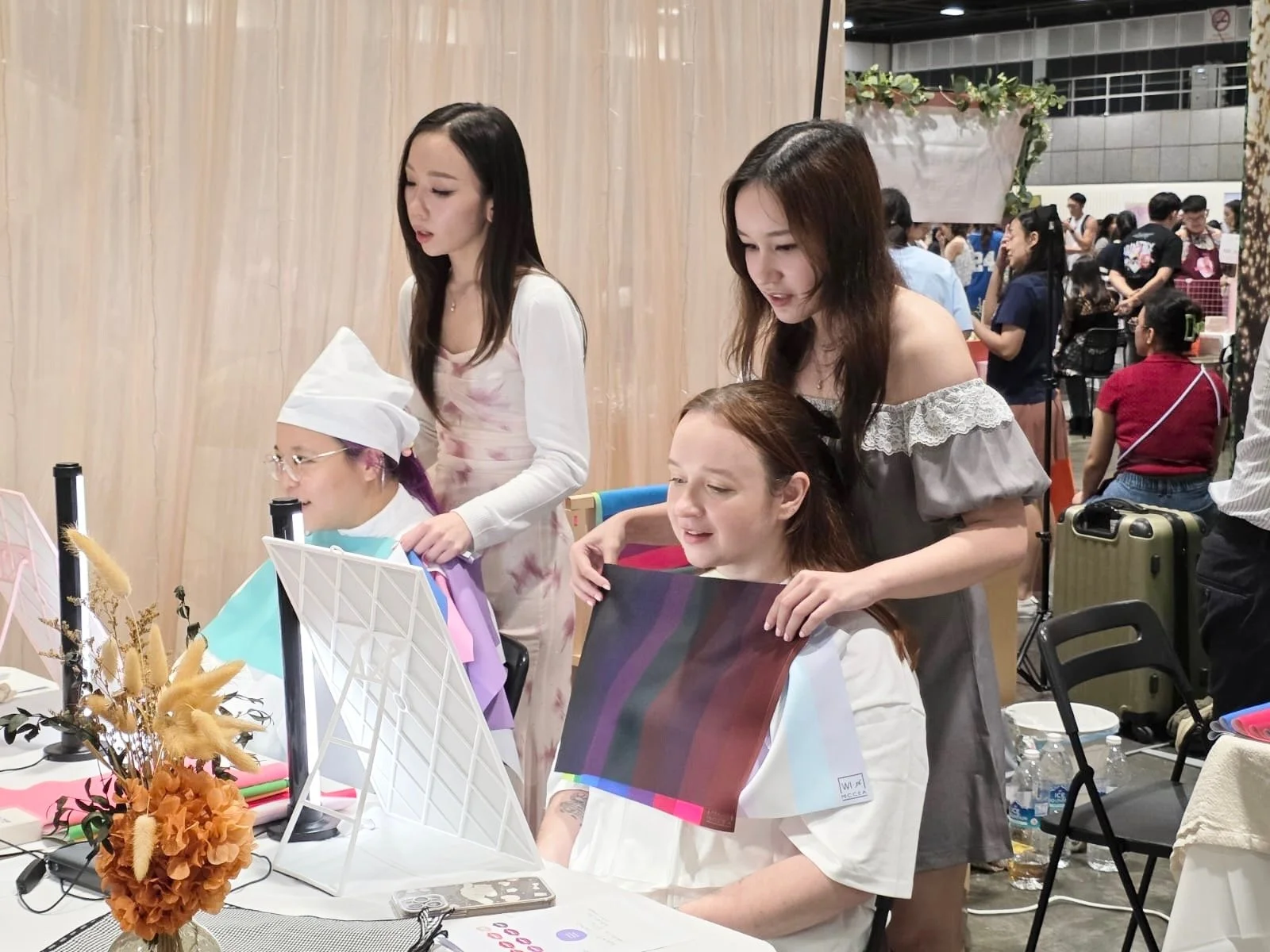 Two women are assisting a seated woman with a hair treatment at a beauty or hair salon convention, with a busy background of people and booths.