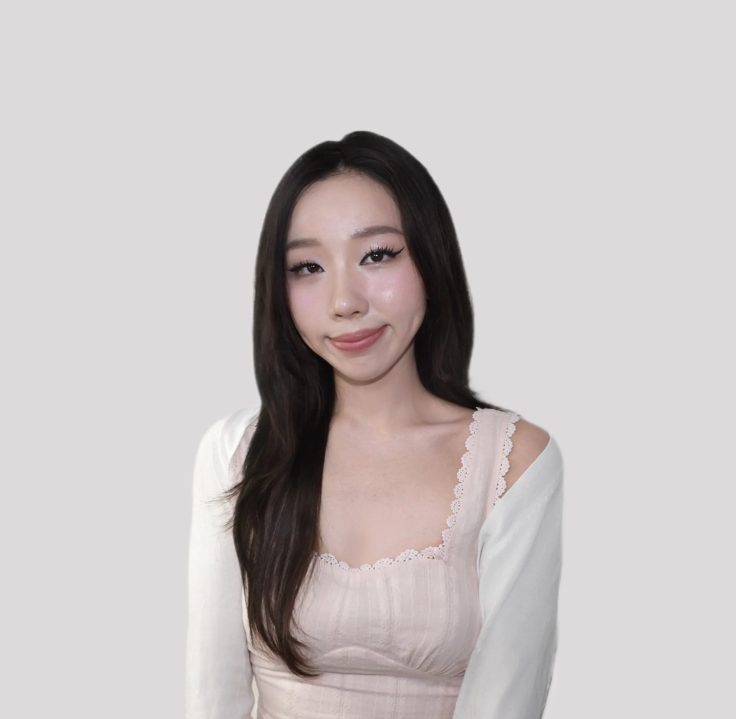 Young woman with long dark hair, wearing a light-colored top with lace detail, standing against a plain light gray background.