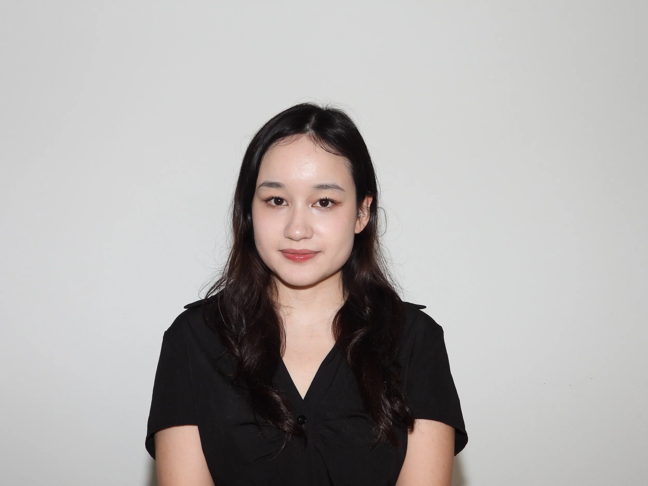 A young woman with wavy dark hair, wearing a black top, standing against a plain white wall.