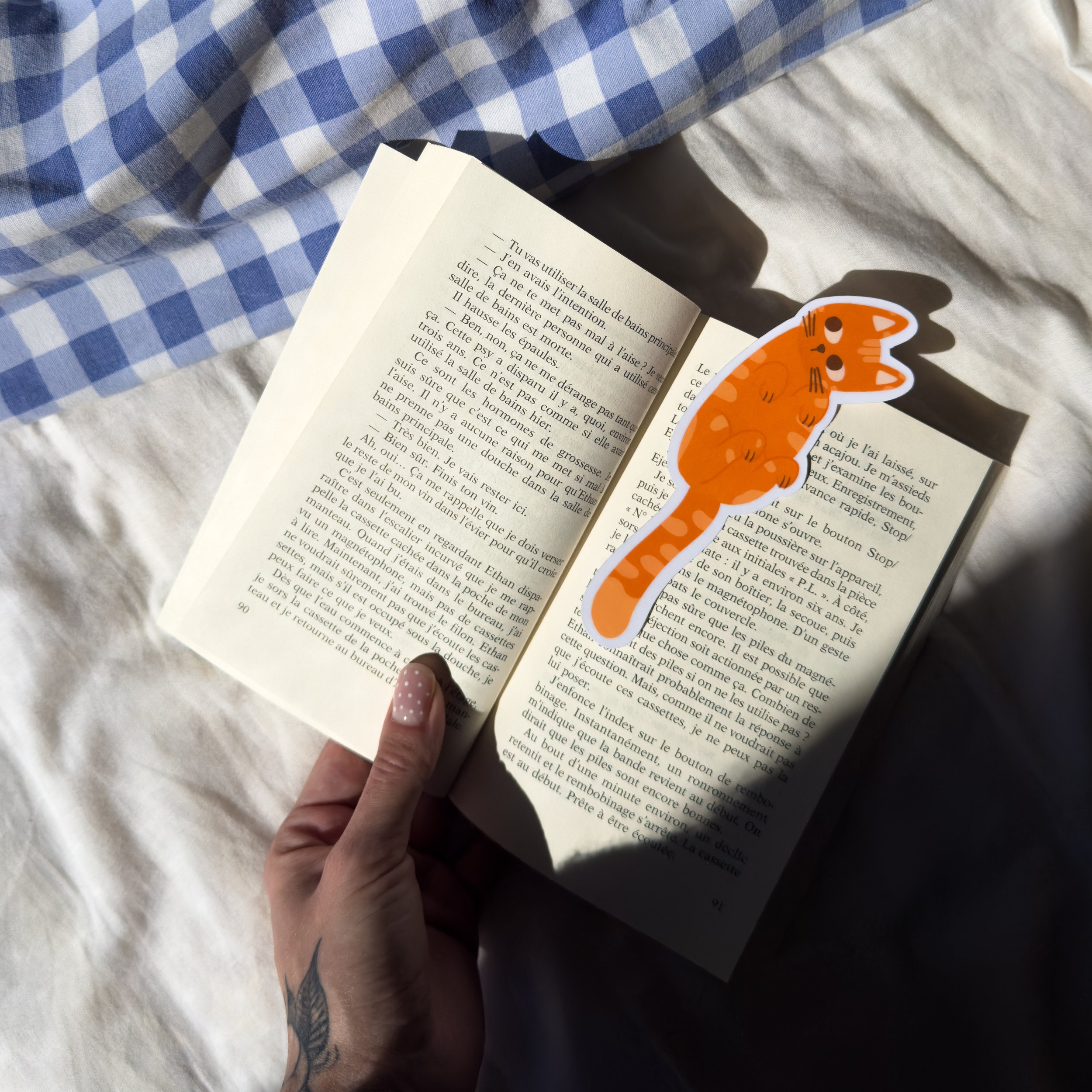 Une main tenant un marque-page en forme de chat orange dans un livre ouvert. Le livre est posé sur une surface recouverte d'un tissu à carreaux bleu et blanc.