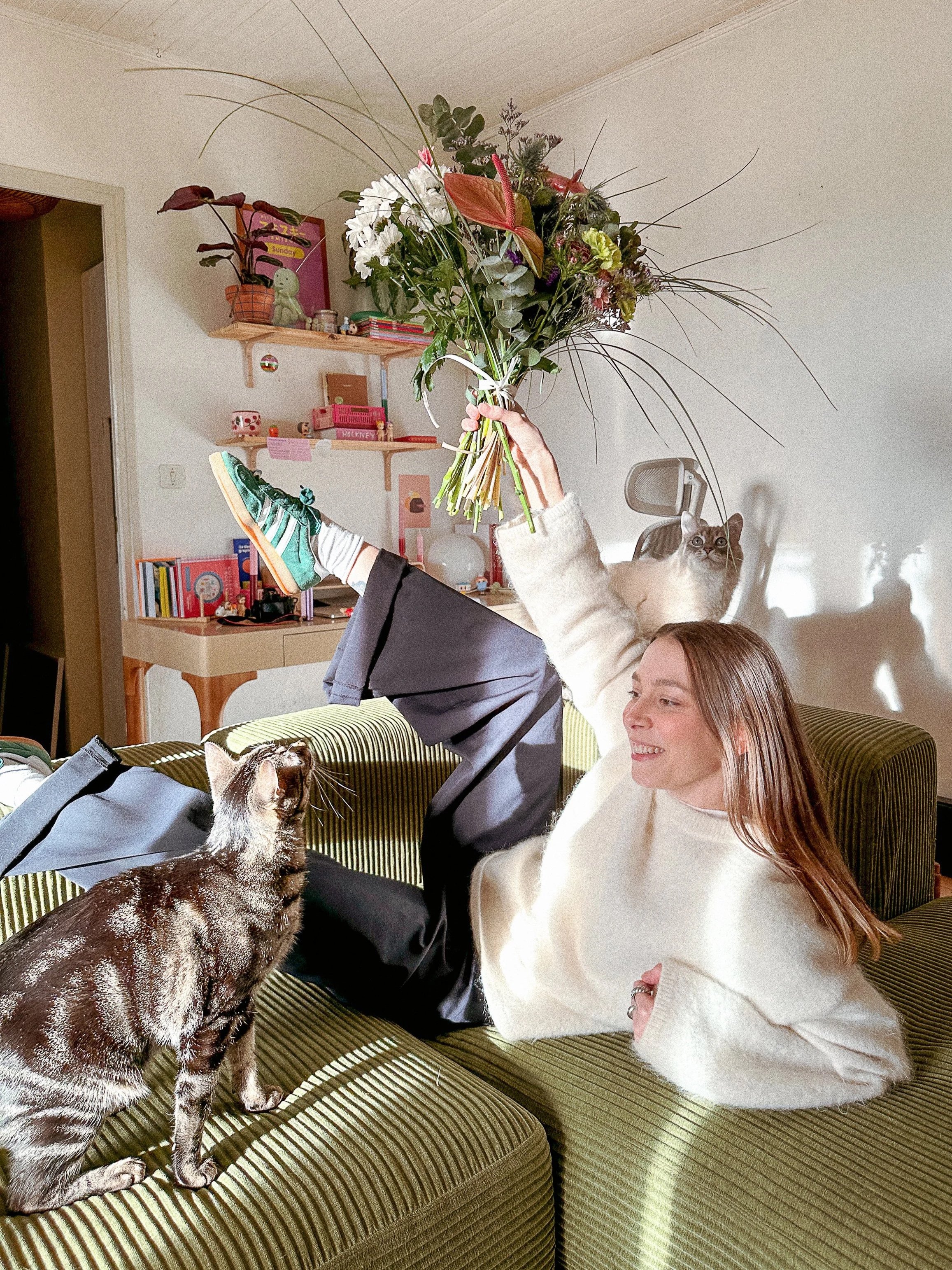 Une femme souriante assise sur un canapé vert, tenant un grand bouquet de fleurs variées, regardant un chat tacheté. La scène se déroule dans une pièce lumineuse avec des étagères décoratives sur le mur et divers objets.
