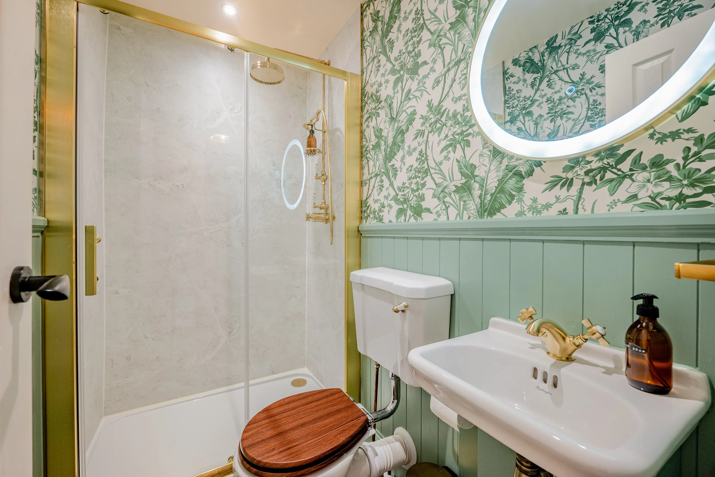 A compact bathroom with a walk-in shower, green botanical wallpaper, a neon-lit oval mirror, a white toilet, and a white sink with gold fixtures and a brown soap dispenser.