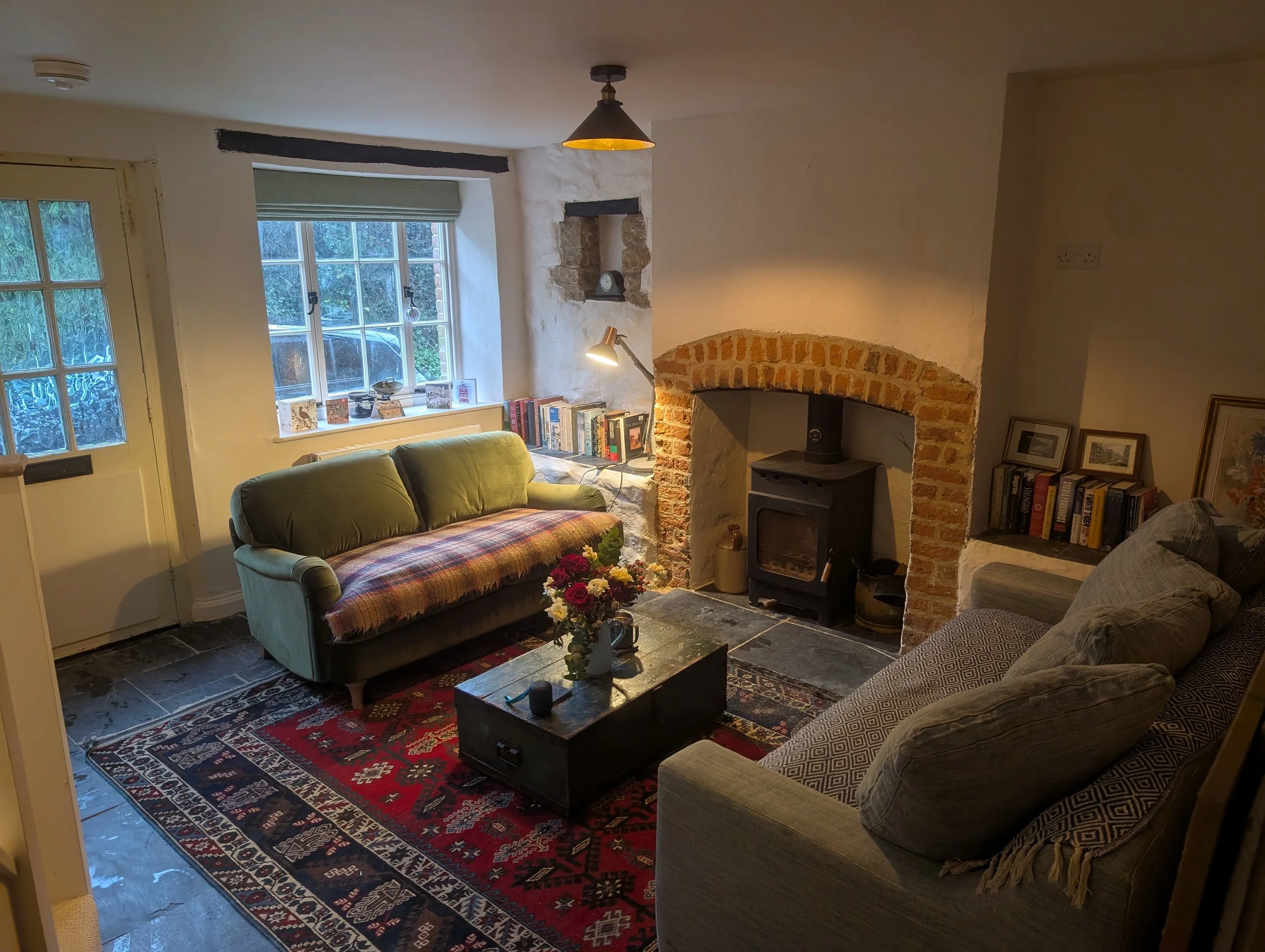 Cozy living room with a green armchair, a wooden coffee table with flowers, a patterned rug, brick and white plaster fireplace, bookshelf, window with green blinds, and framed artwork.