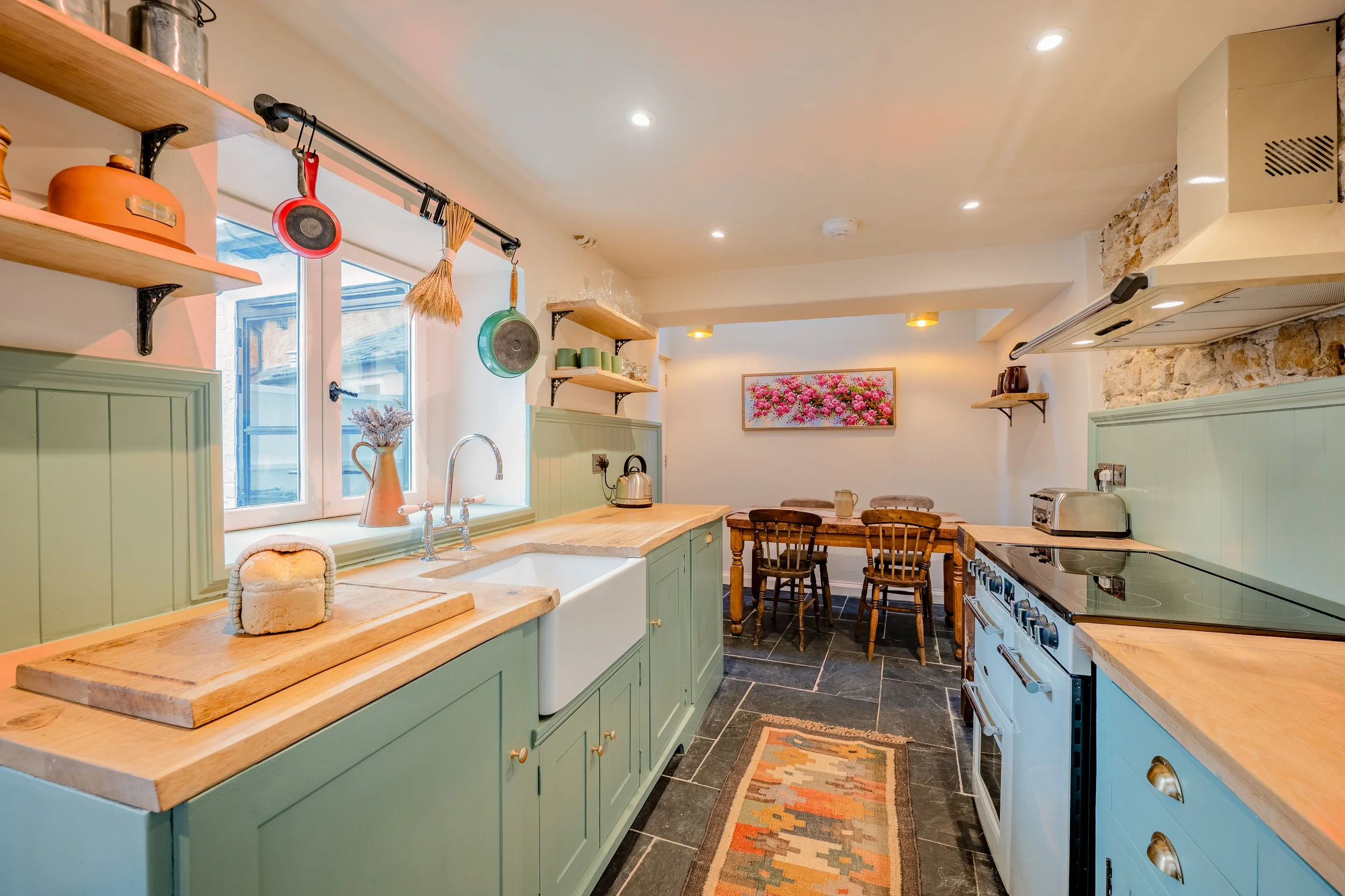 A cozy kitchen with light green cabinets, a wooden countertop, hanging pots and pans, a window with a view outside, a kitchen sink, a small dining table with chairs, and wall shelves with dishes and decorative items.