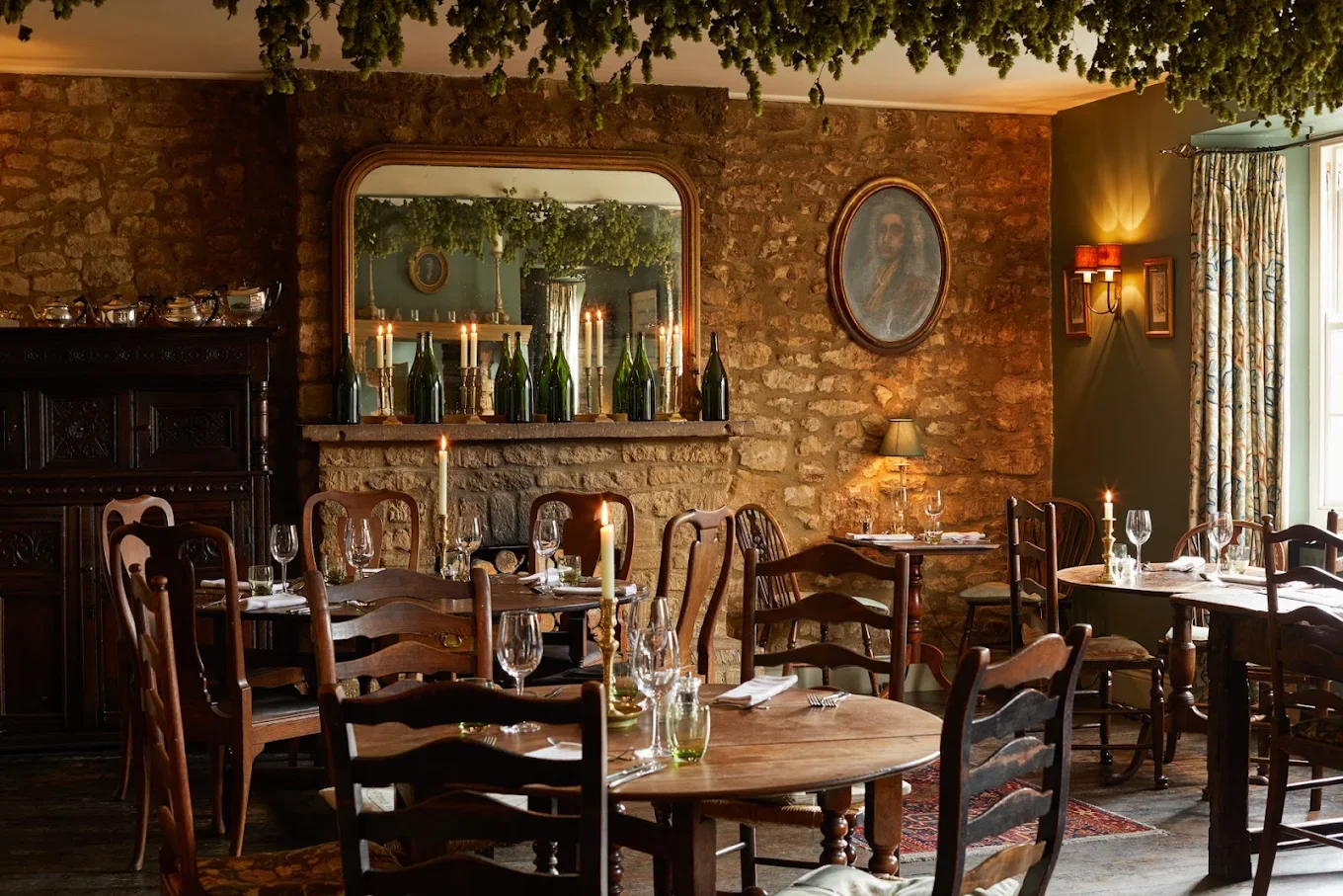 Cozy rustic restaurant with stone wall, wooden tables and chairs, candlelit ambiance, bottles on a shelf, and warm lighting.