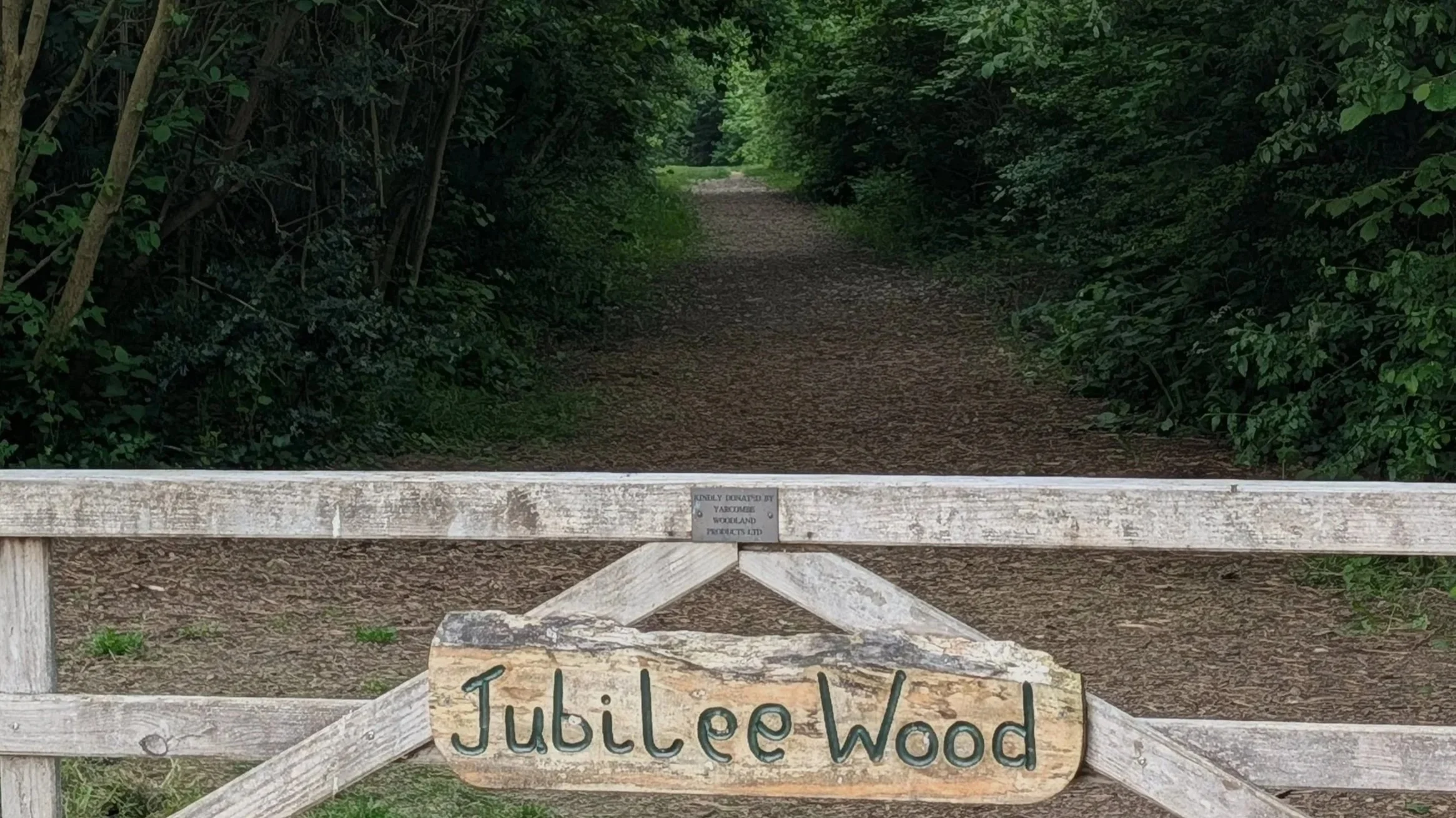 A wooden gate with a sign that reads "Jubilee Wood" blocking a dirt path through a lush green wooded area.