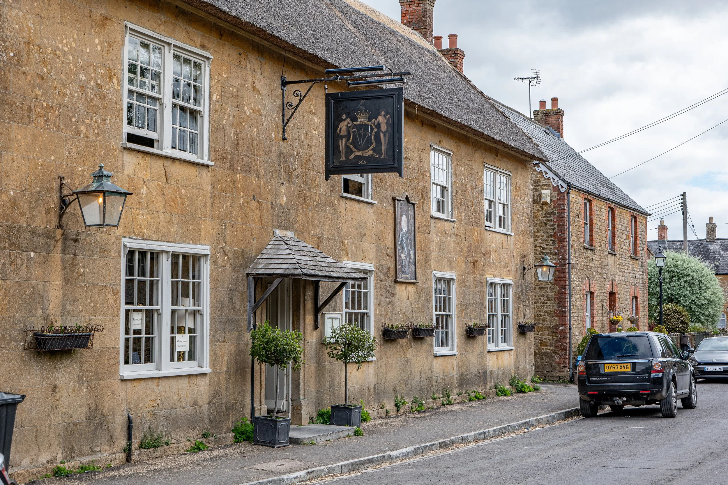 A street view of a building with a stone facade and white-framed windows. There are black planters with plants beneath some windows, a small covered entrance porch, and a vintage lantern on the wall. A black sign with painted figures hangs above the street, and multiple cars are parked along the curb, including a Land Rover and an Audi. The sky is overcast.