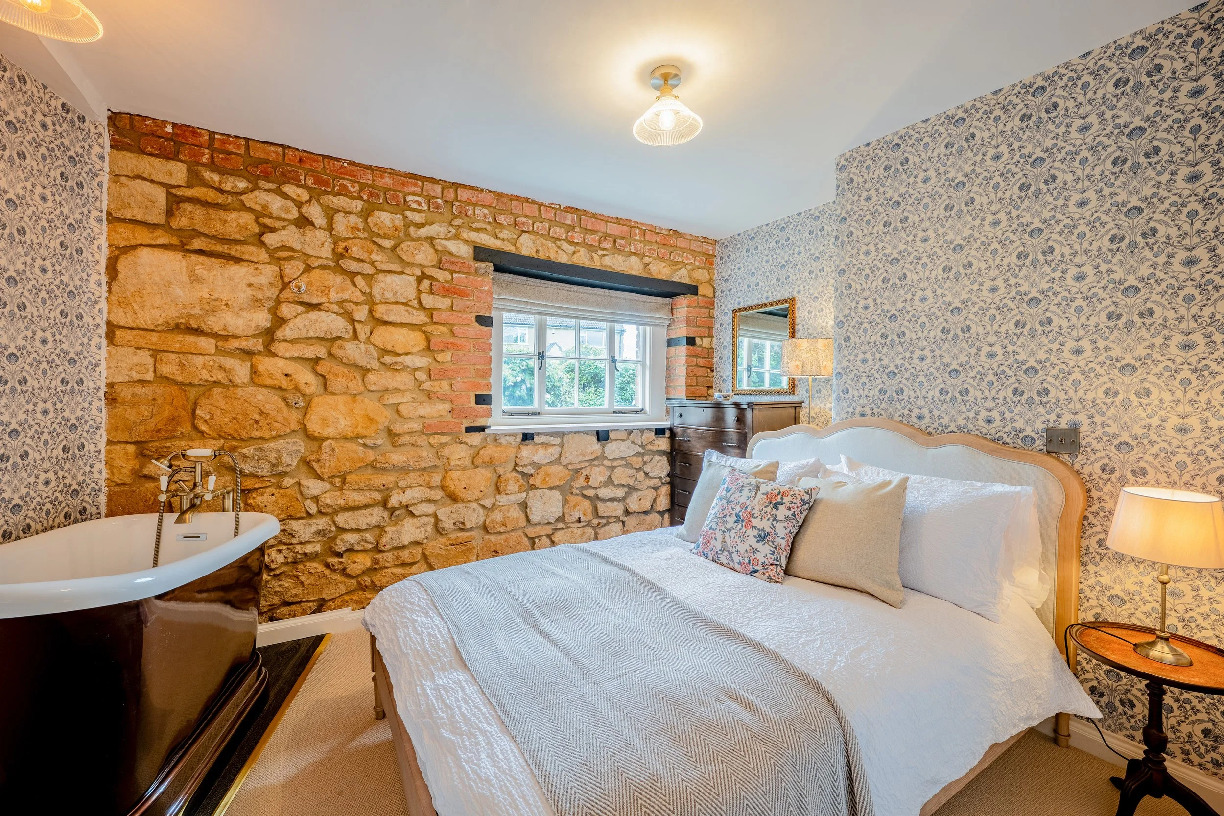 A cozy bedroom featuring a stone brick accent wall, a window with a Roman shade, and floral wallpaper.