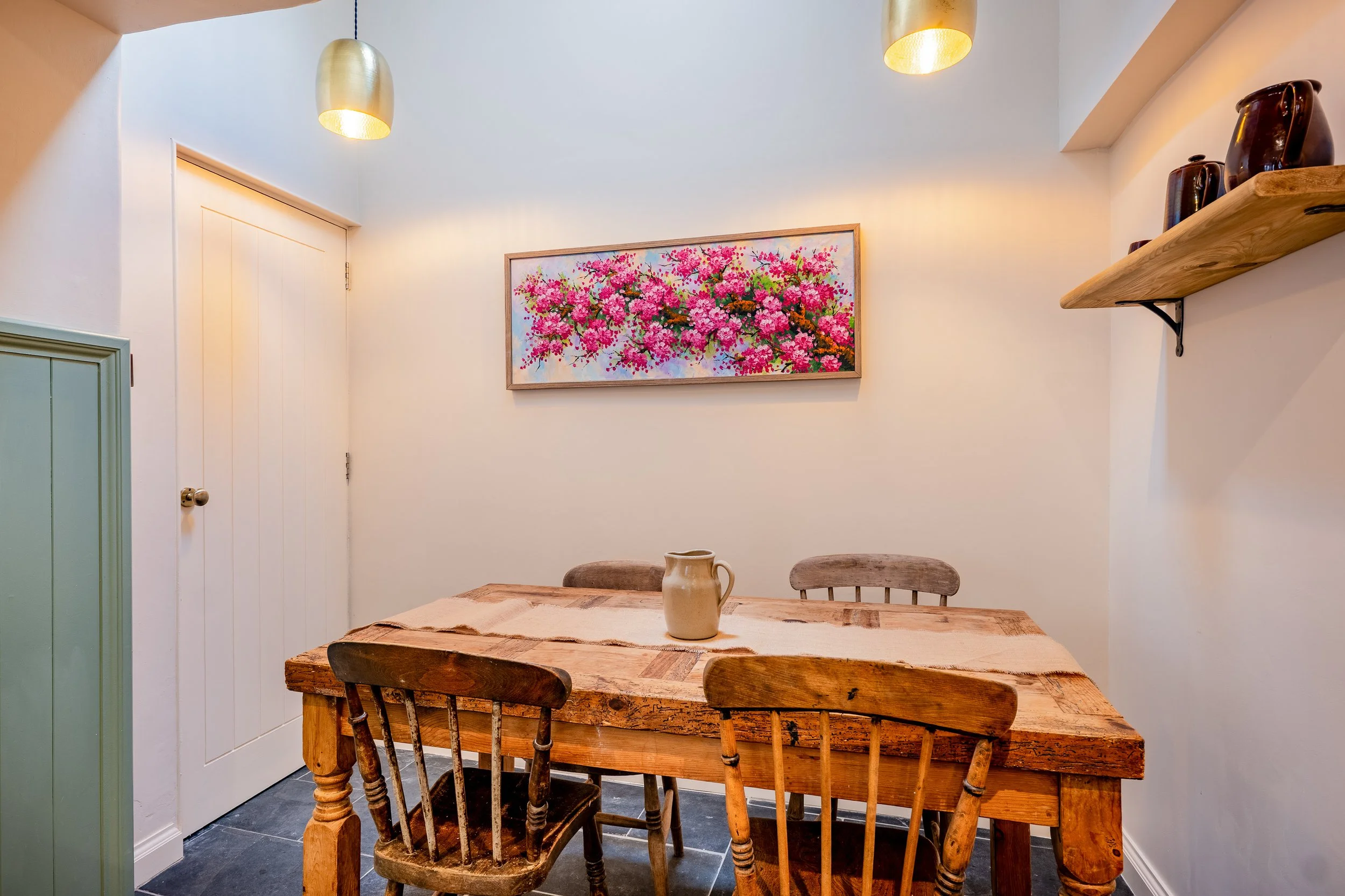 A rustic wooden dining table with a beige pitcher and four wooden chairs in a small dining nook. The walls are painted white, with a floral painting featuring pink blossoms hanging on one wall, and a wooden shelf with dark brown vases on the right.