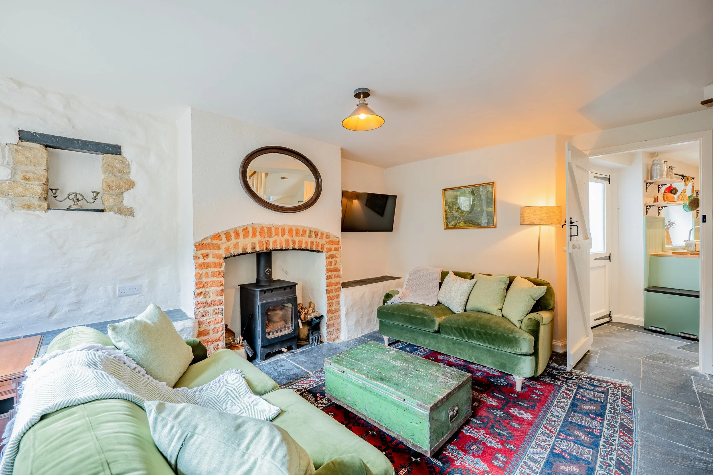 Living room with a green sofa, wood stove beneath a brick fireplace, wall mirror, mounted TV, framed picture, floor lamp, and a colorful area rug.