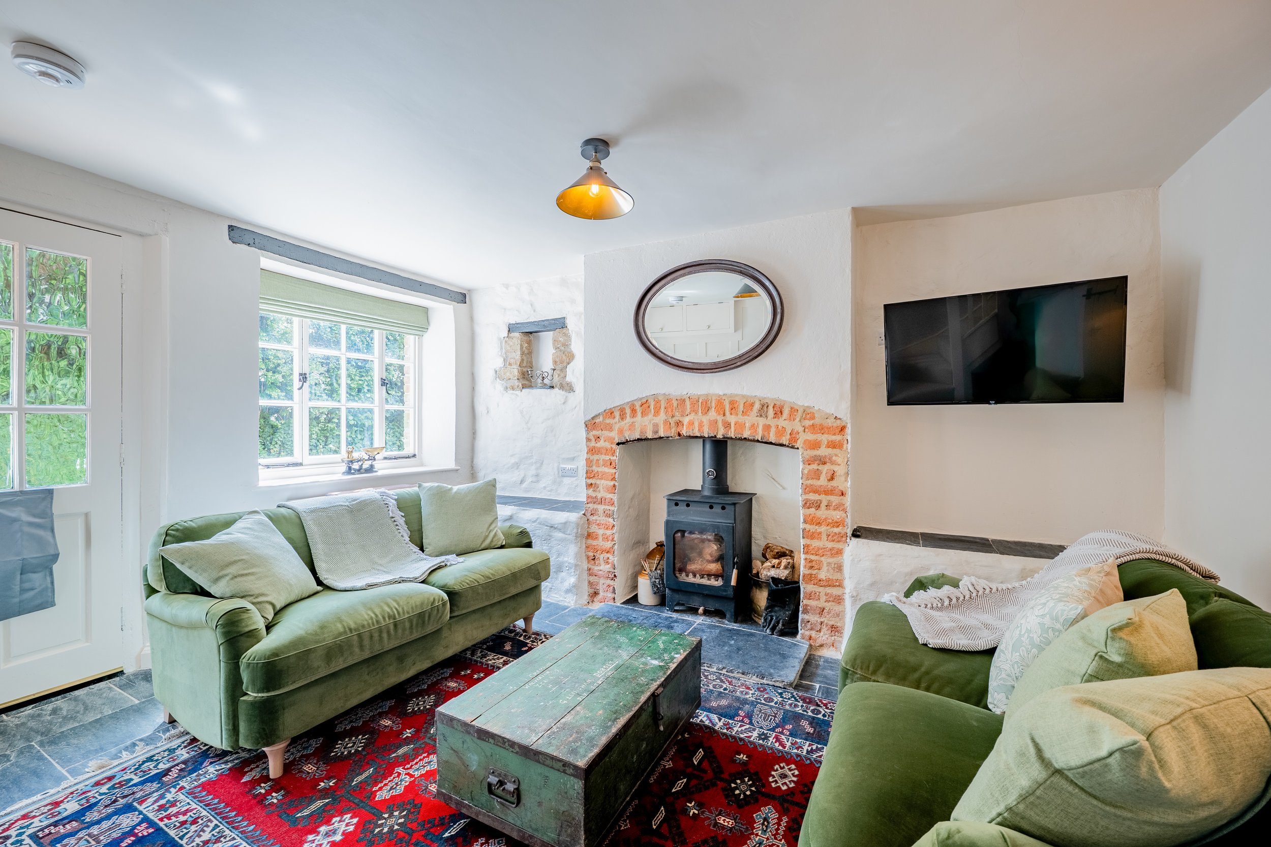 Cozy living room with green sofas, a wood-burning stove with a brick surround, a window with blinds, a decorative mirror, a flat-screen TV, and a colorful oriental rug.