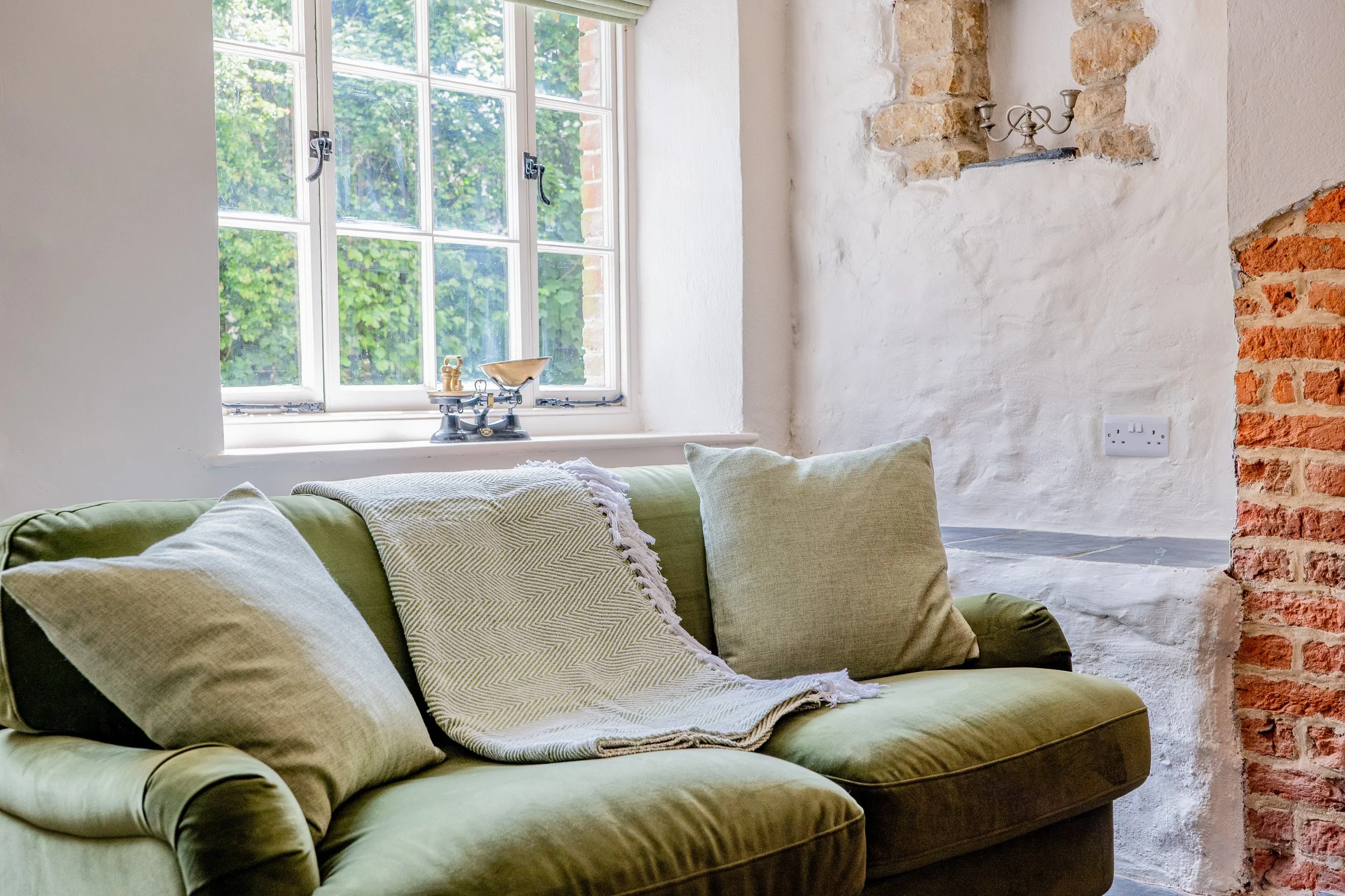 A cozy living room with a green velvet sofa, light-colored pillows, and a blanket. A large window with white framing shows greenery outside. There is a brick wall and a white painted wall with an electrical outlet. Decorative objects on the windowsil
