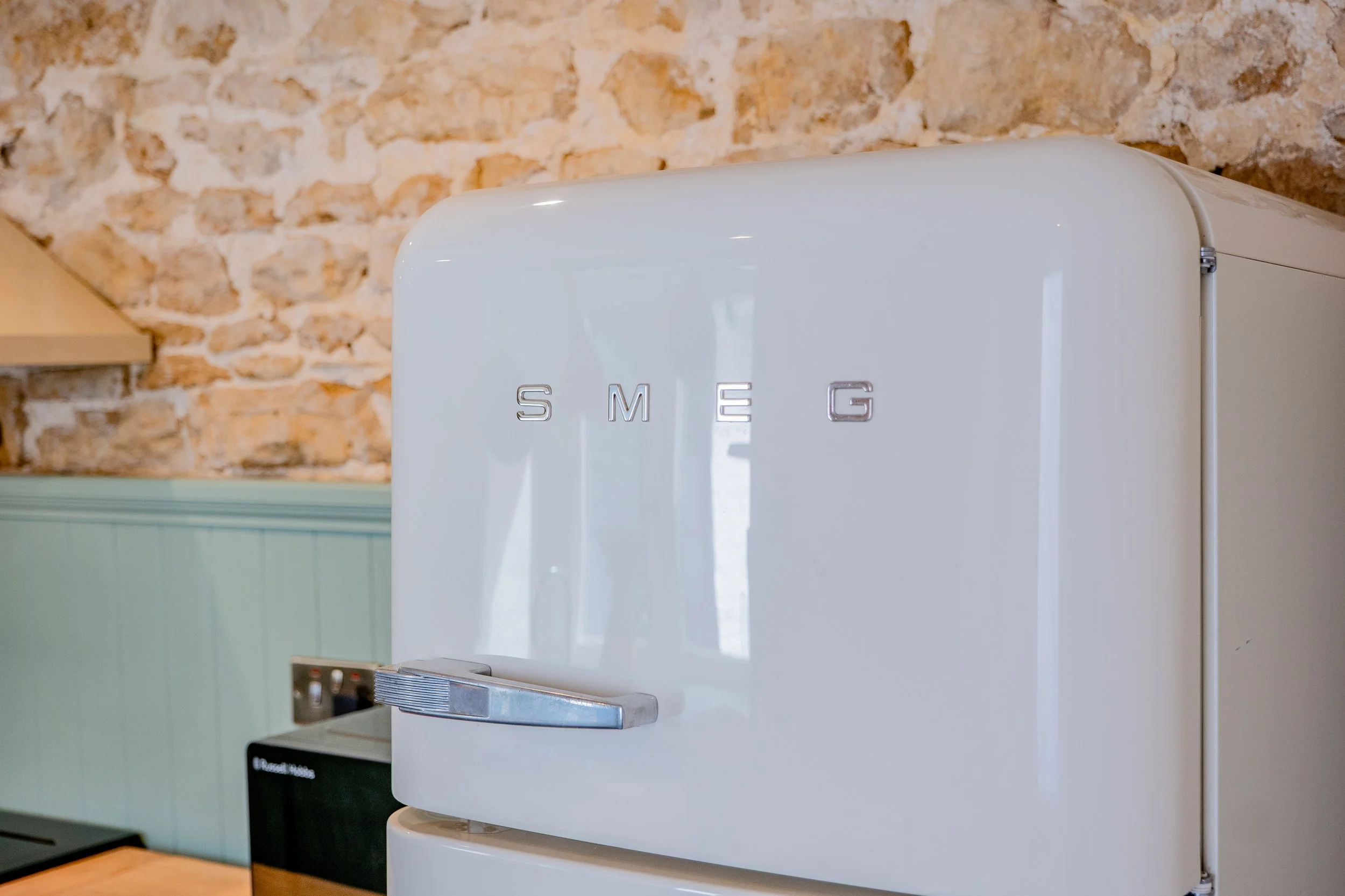 A white SMEG refrigerator against a stone brick wall, with a metallic handle and silver brand logo.