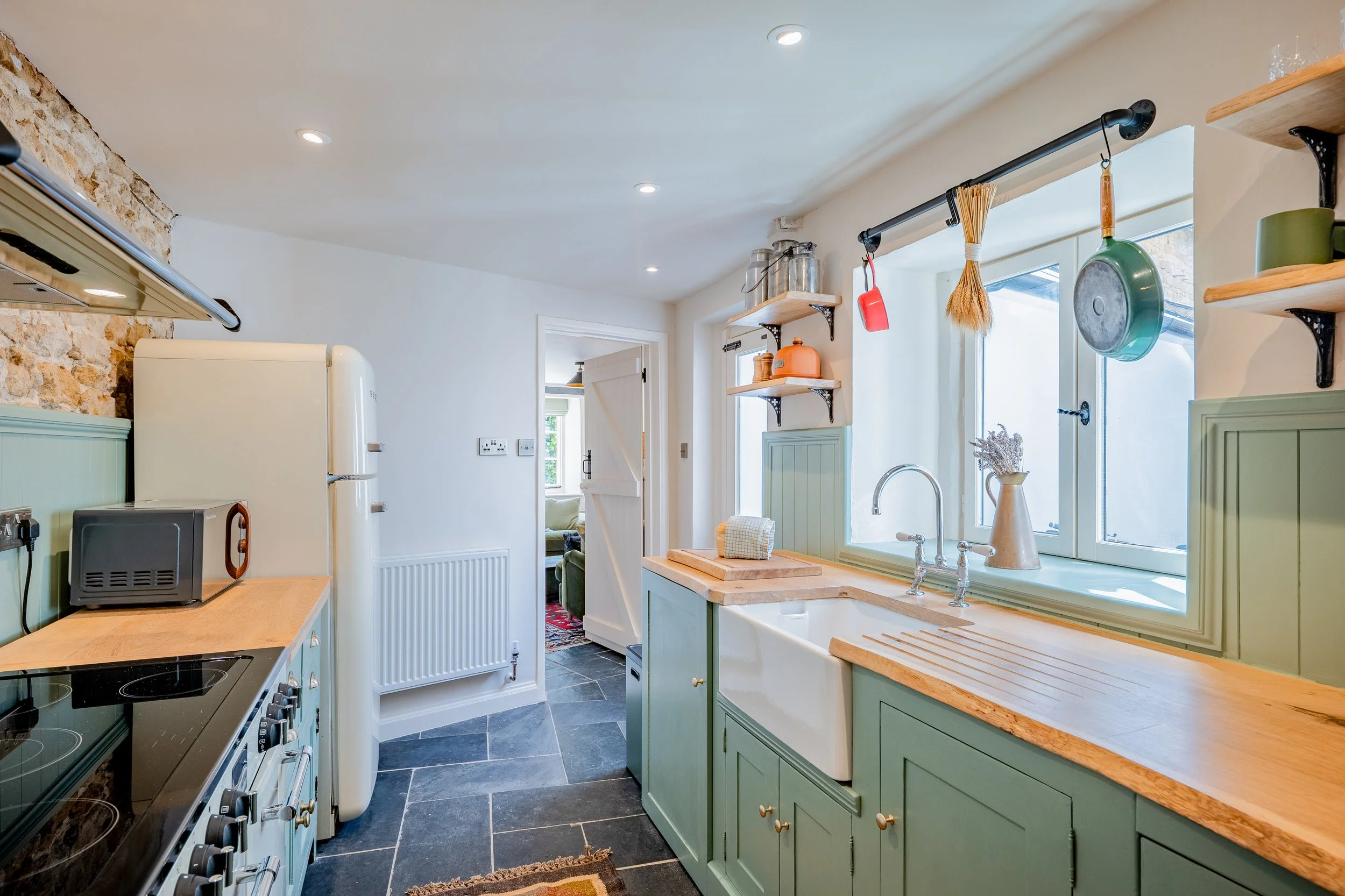 A cozy kitchen with light green cabinets, a wooden countertop, a white farmhouse sink, hanging pots and utensils, and a view into a living room with a sofa.