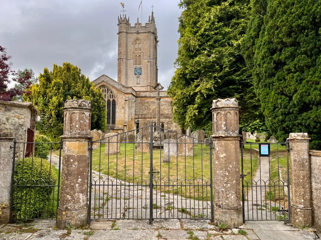 HintonSt George church with a tall tower, surrounded by stone walls and green trees. The church is viewed behind a black wrought iron gate.