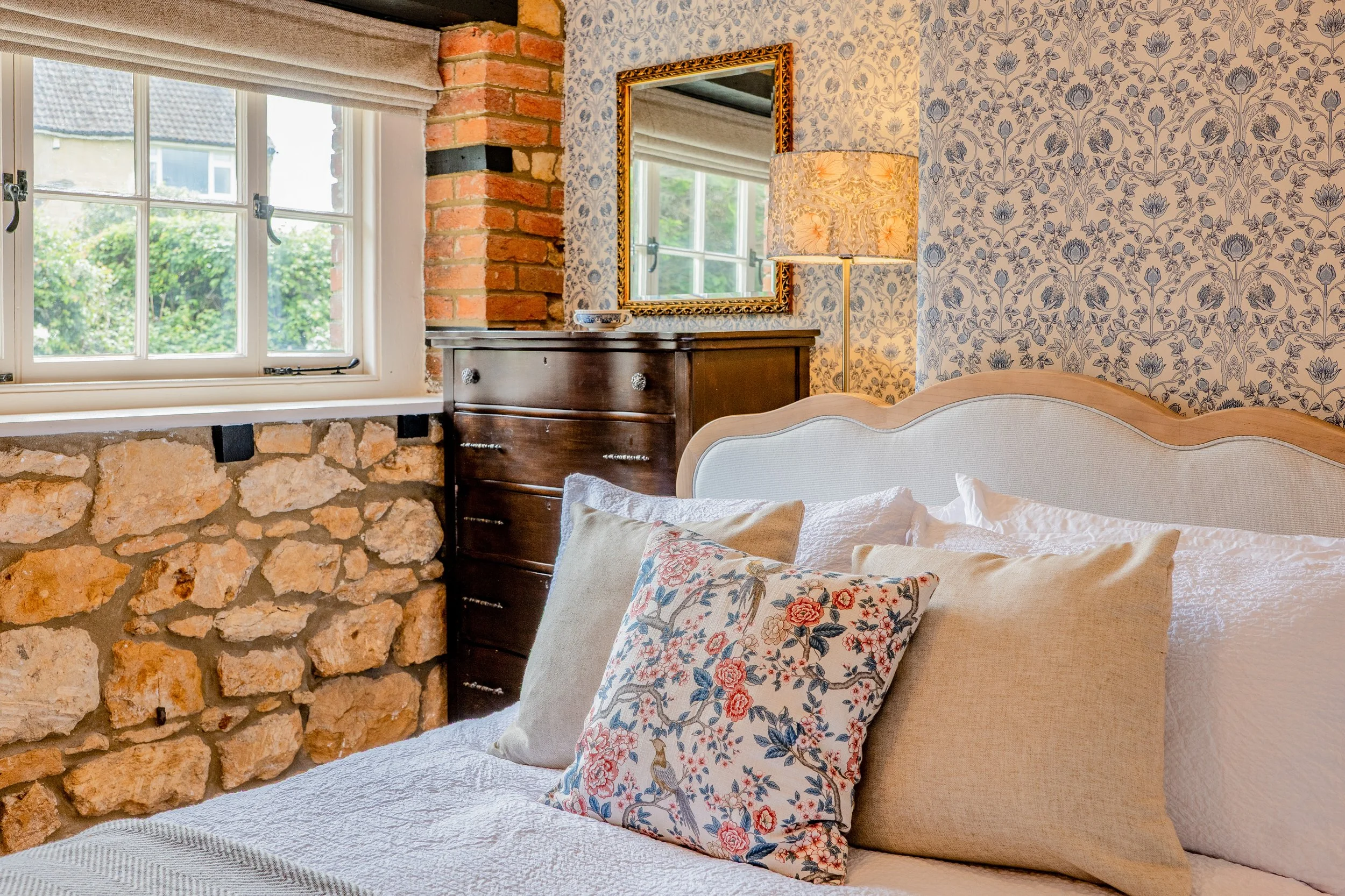 Close-up view of a bedroom with a window, brick and stone walls, floral wallpaper, a mirror, a lamp, and a bed with pillows.