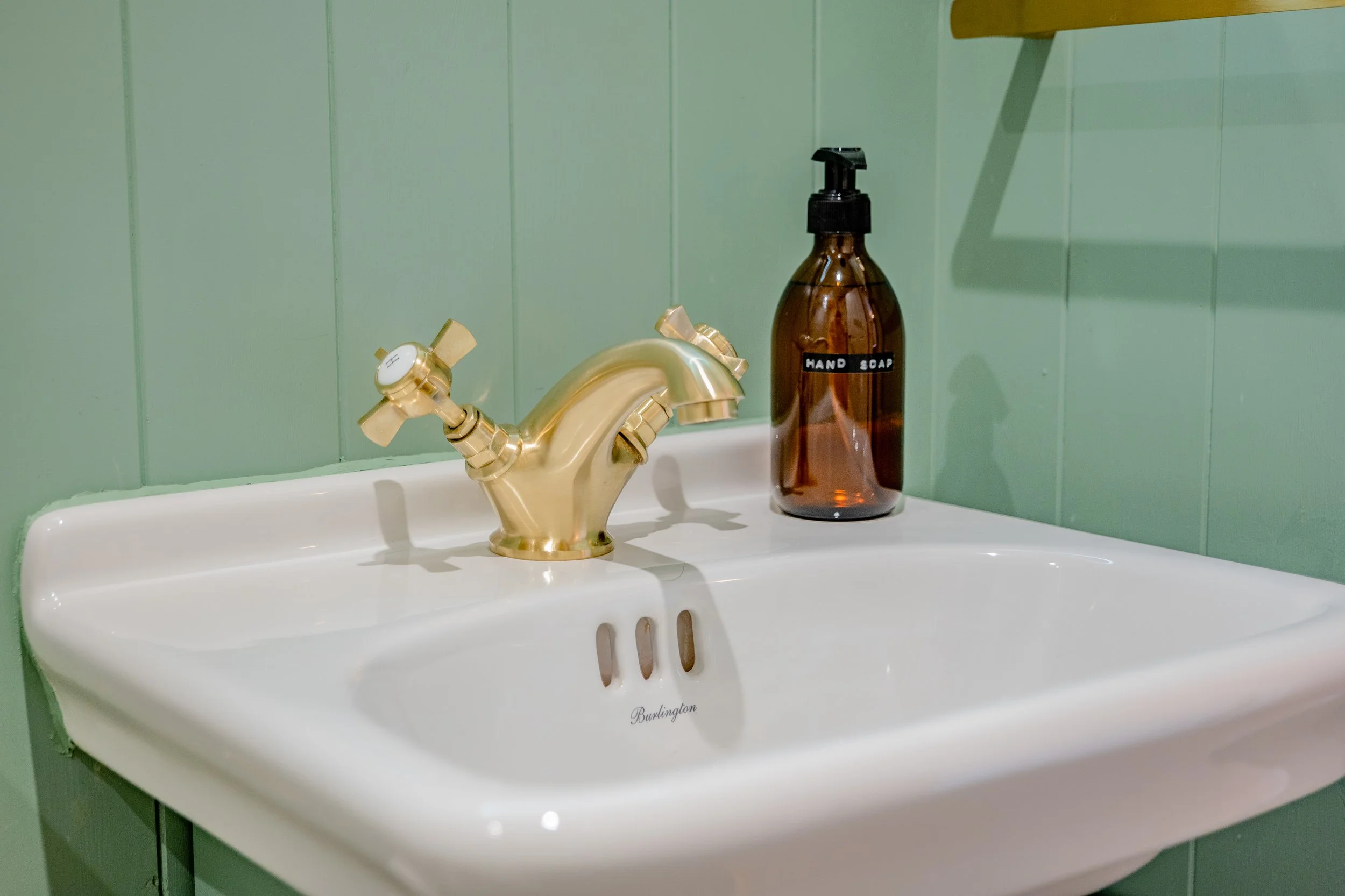 A white porcelain bathroom sink with a gold-colored faucet, an amber soap bottle labeled "Hand Soap," against a light green tiled wall.