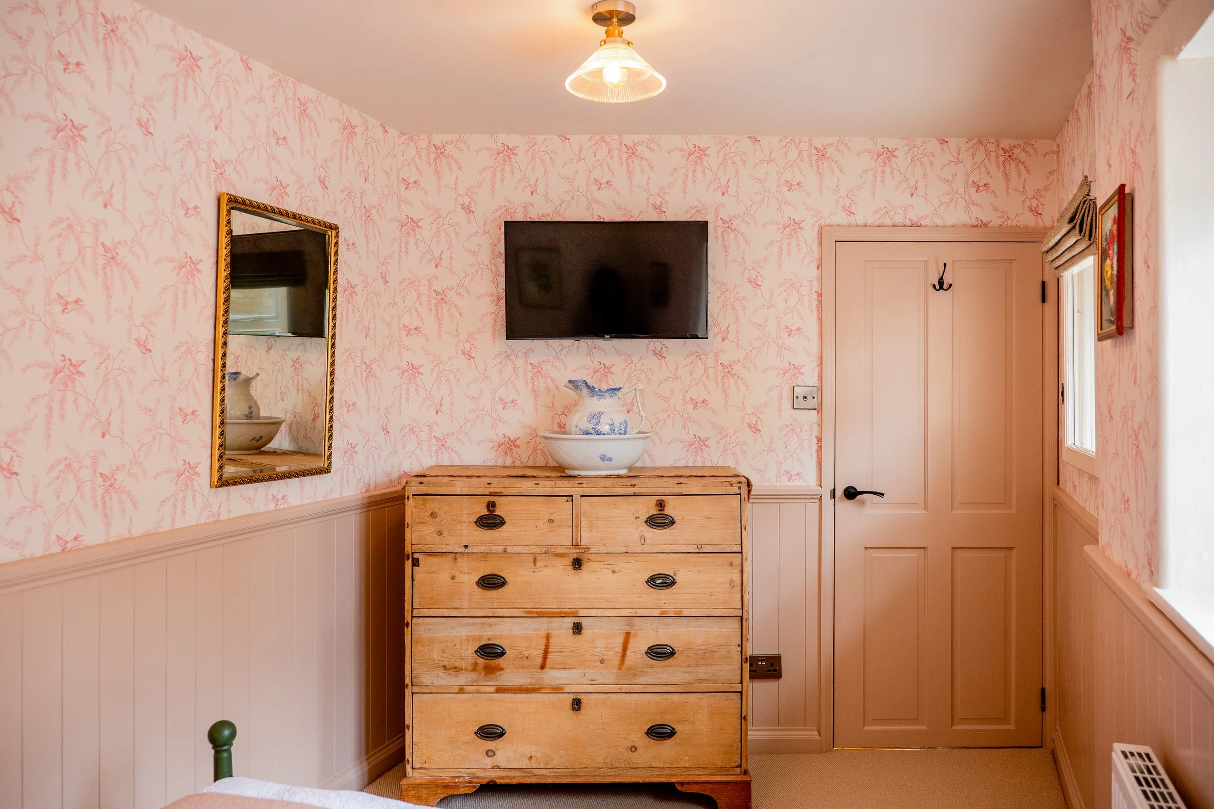 A vintage wooden dresser with black metal handles, topped with a large decorative white and blue porcelain bowl. Behind the dresser is a pink wallpapered wall with a pattern of pink plants and birds. To the left, a framed mirror hangs on the wall. To