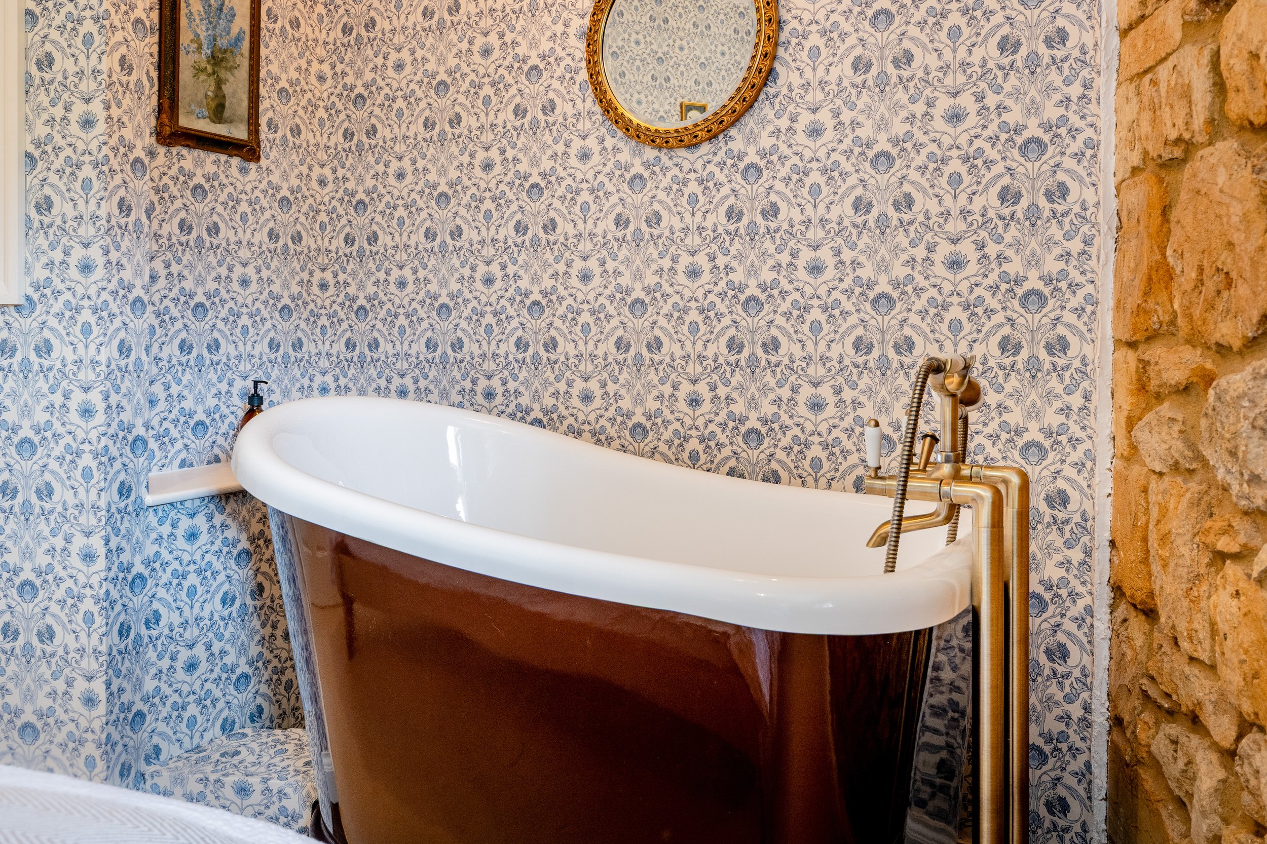 A vintage-style rolltop bath , decorative blue and white floral wallpaper, a framed floral painting, a round gold-framed mirror, and exposed brick wall.