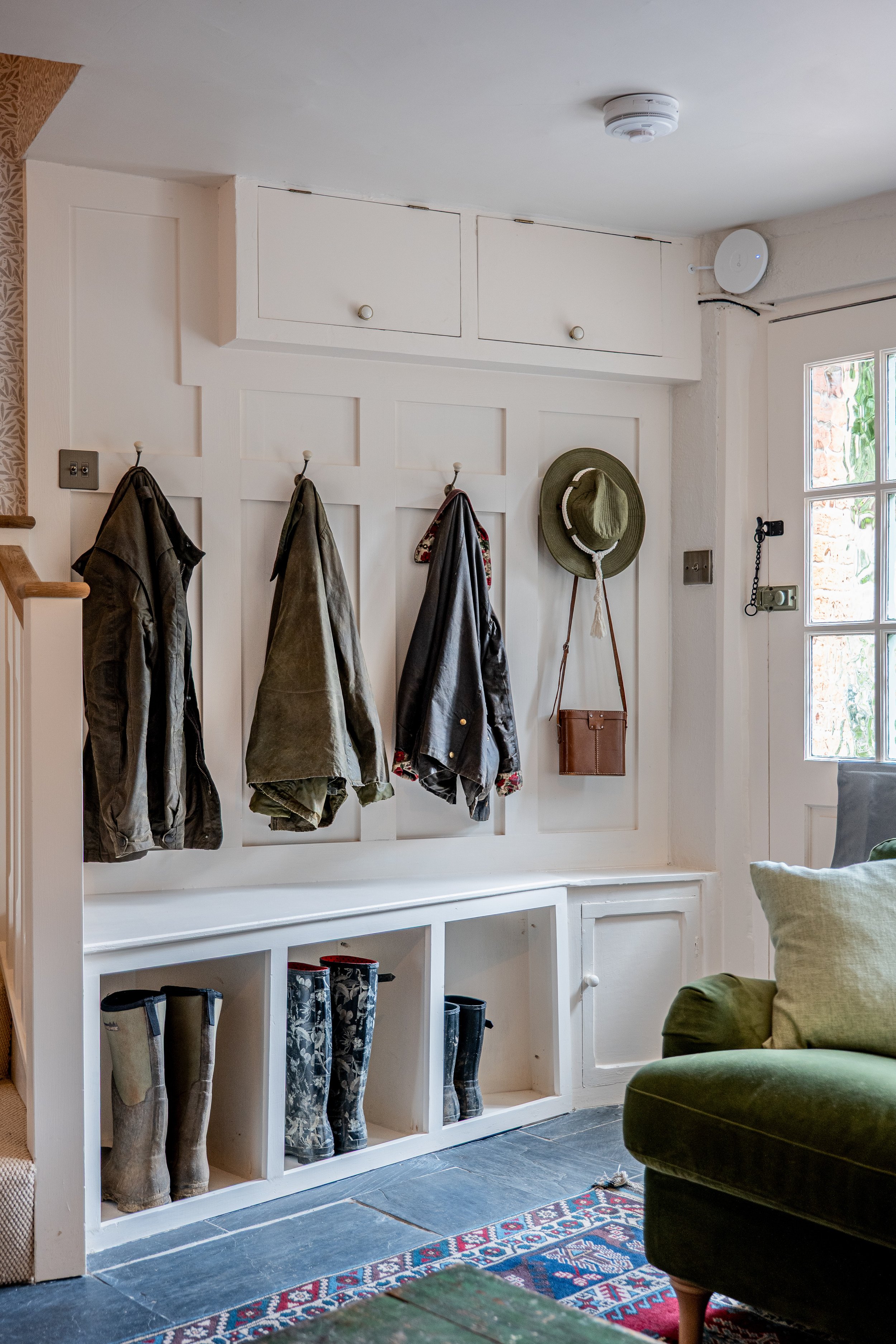 Coat hooks on wall with jackets hanging, hats and a bag displayed, black rain boots in cubbies, and part of a green chair in a cozy, rustic entryway.