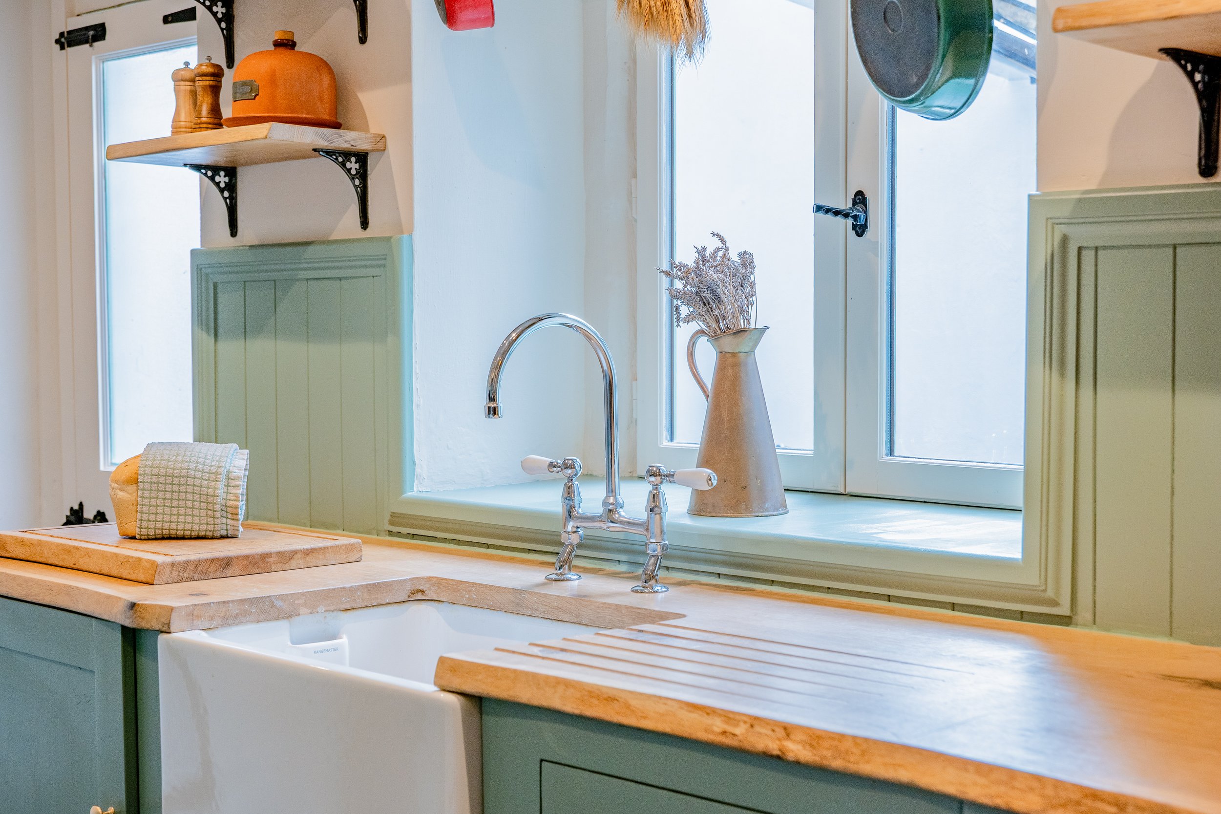 A kitchen window with a beige ceramic pitcher holding dried flowers, a green cabinet below, and a wooden cutting board with a cloth on it on the countertop. There is a double sink with a chrome faucet.