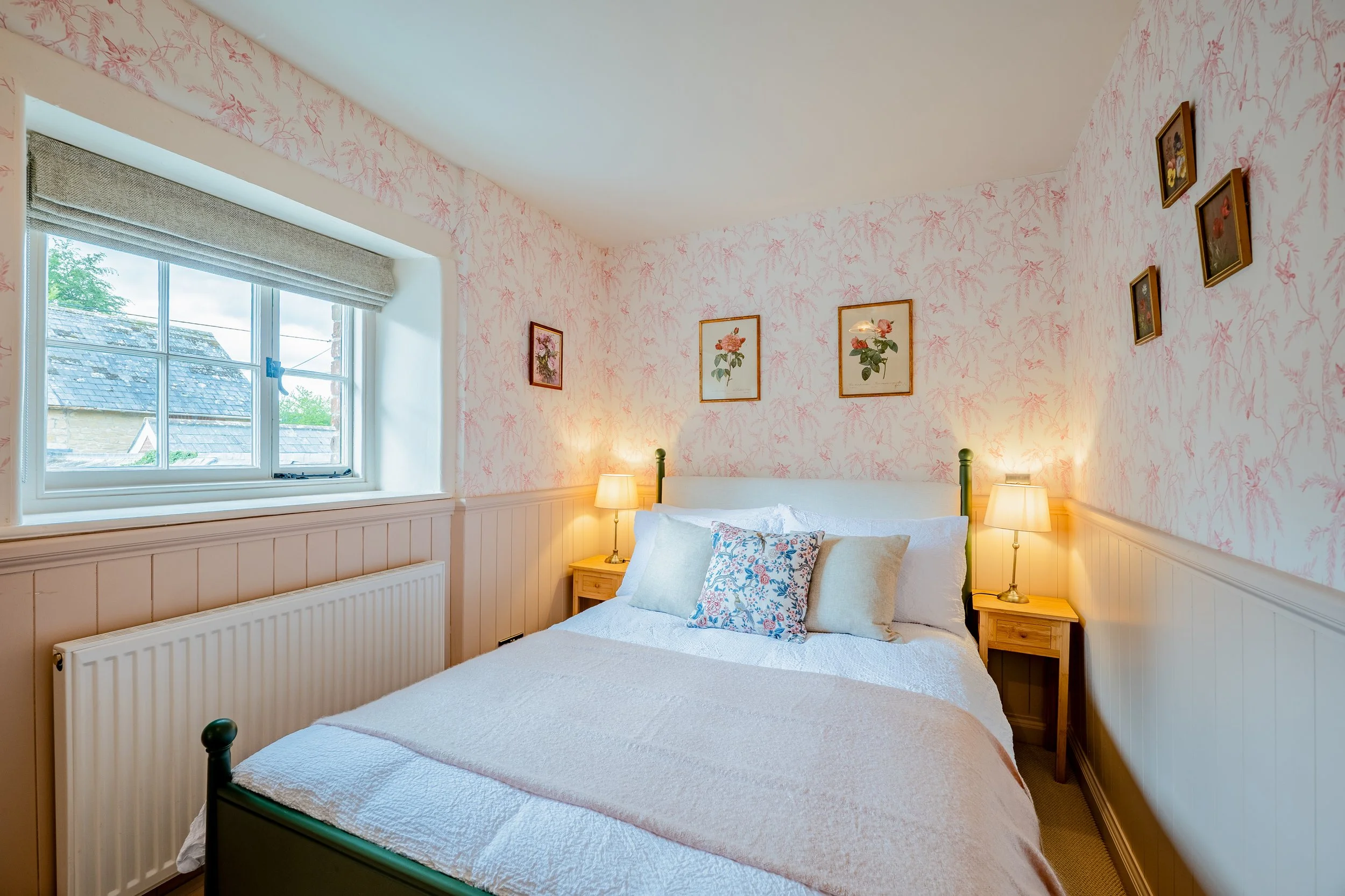 A cozy bedroom with a window, floral wallpaper, a double bed with white bedding and floral pillows, two bedside tables with lamps, and framed botanical prints on the wall.