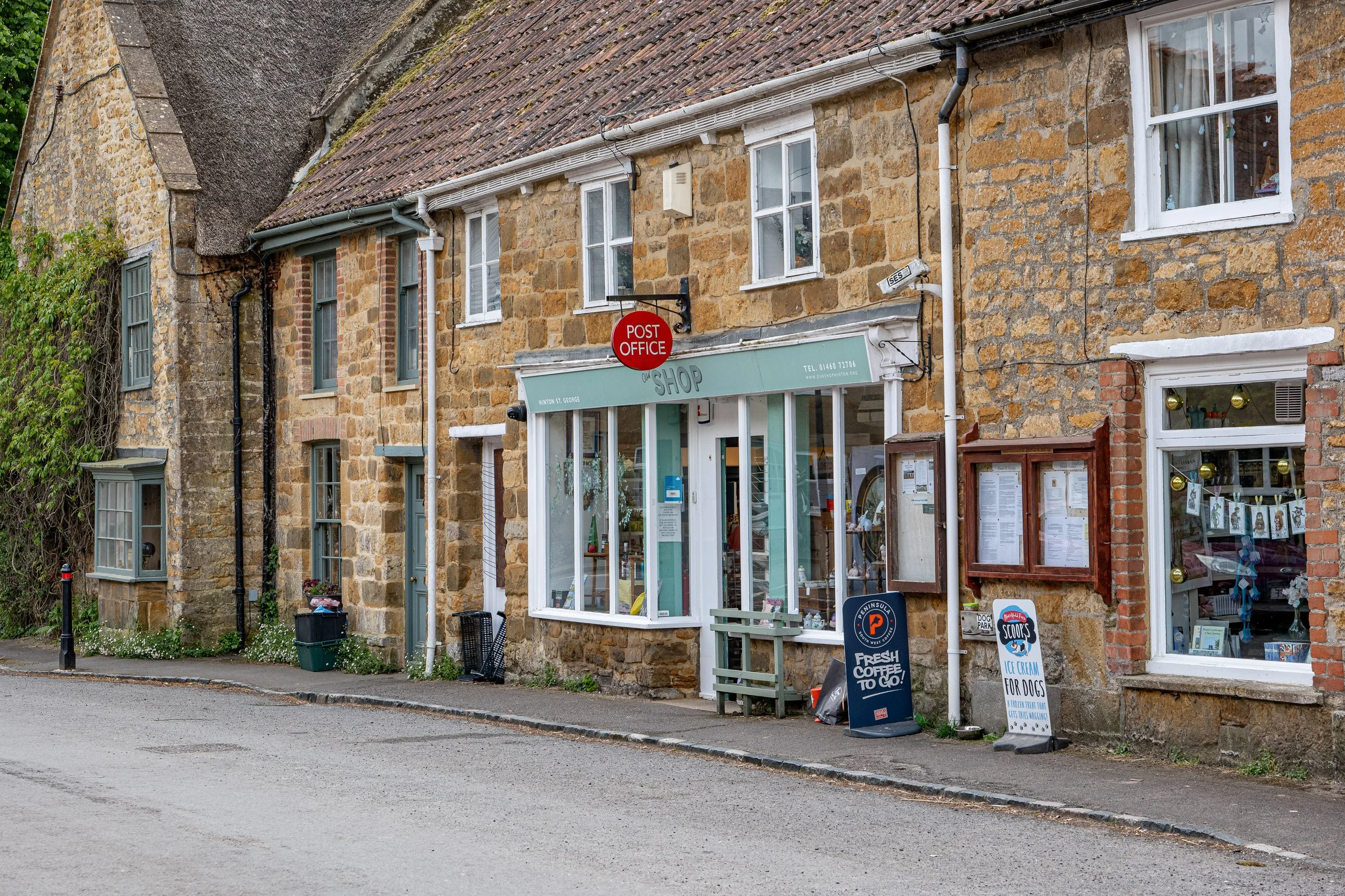 A street view of Hinton St George village shop on the ground floor, featuring a green awning, large windows, and signs advertising coffee, ice cream, and a post office in a small town.