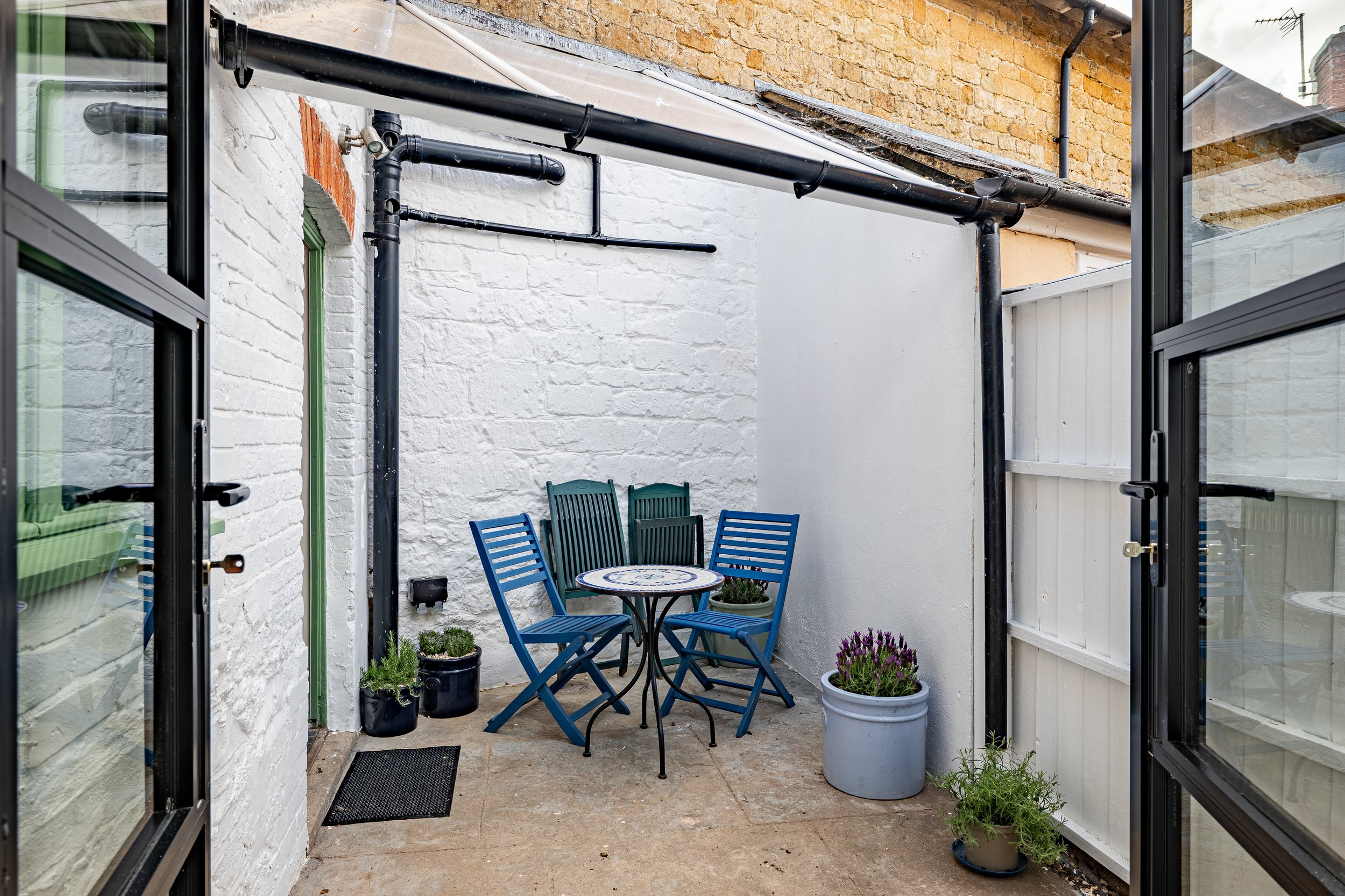 An outdoor patio viewed through open glass doors, with a small round metal table and four blue chairs, potted plants, and a white brick wall.