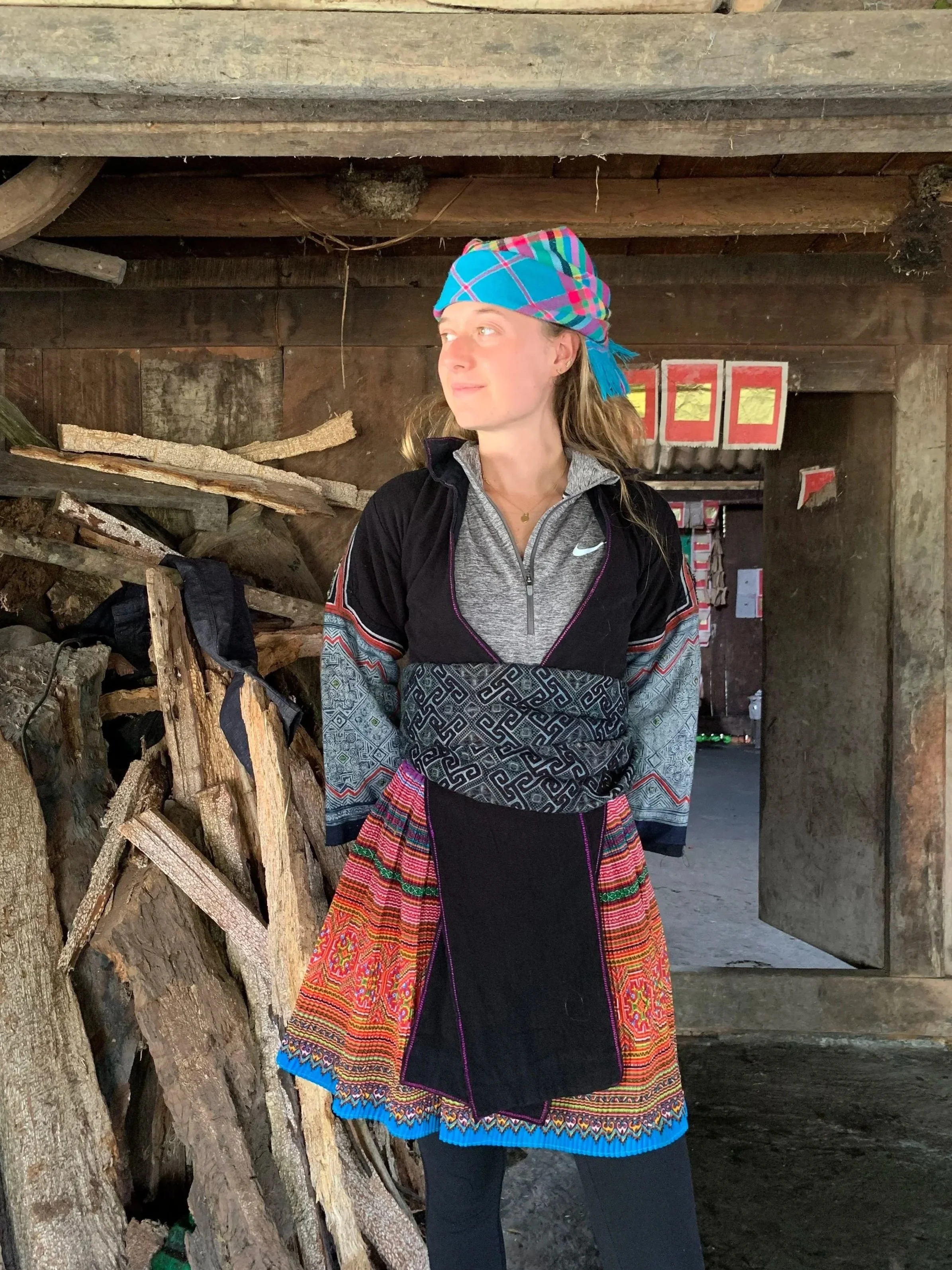 A woman standing inside a rustic wooden shed, wearing a colorful headscarf, a zip-up quarter-zip jacket, and a vibrant, woven apron, with a pile of firewood behind her.