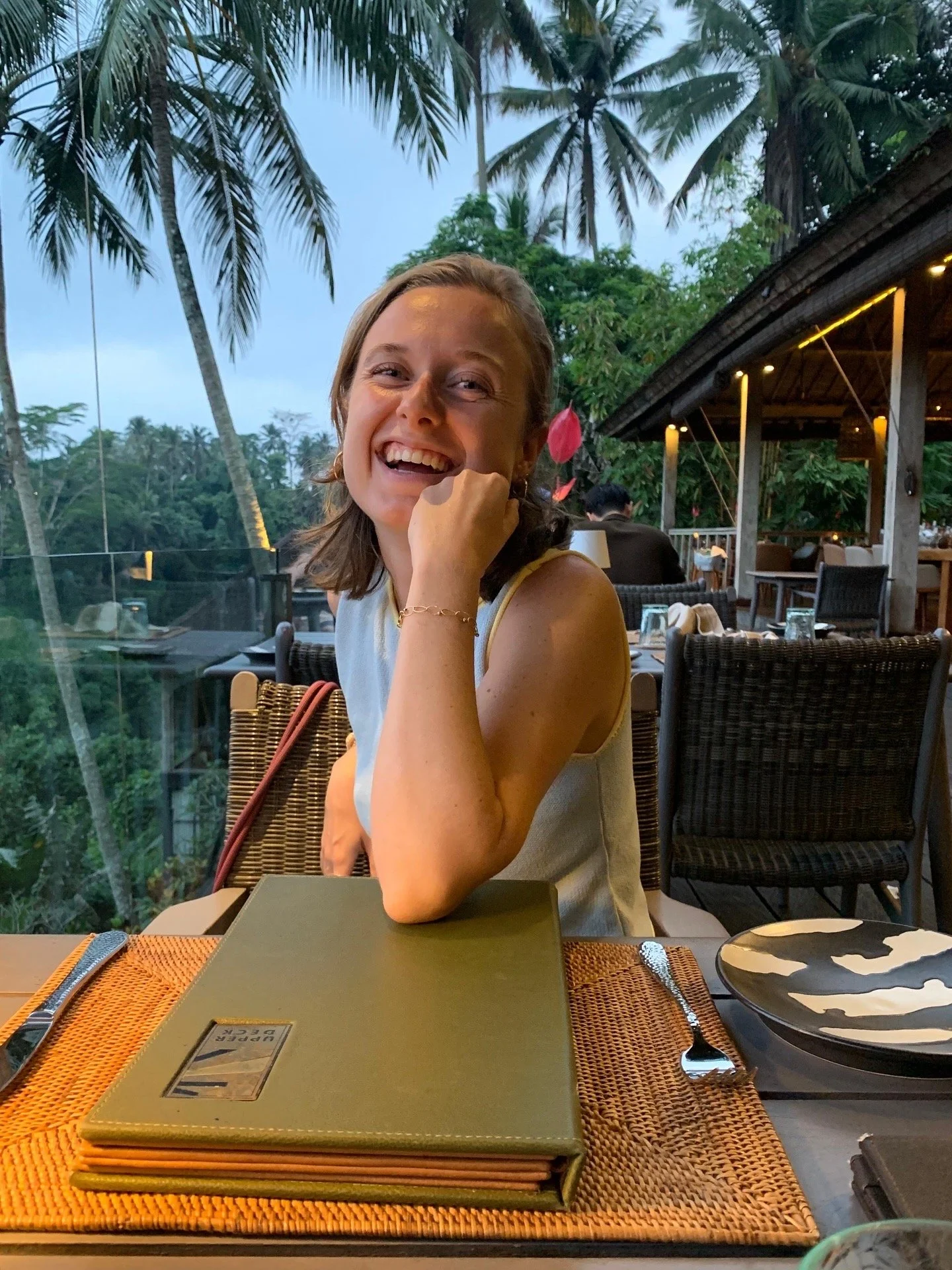 A woman sitting at a table outdoors, smiling, with a tropical background of palm trees, a restaurant with string lights, and a glass railing.