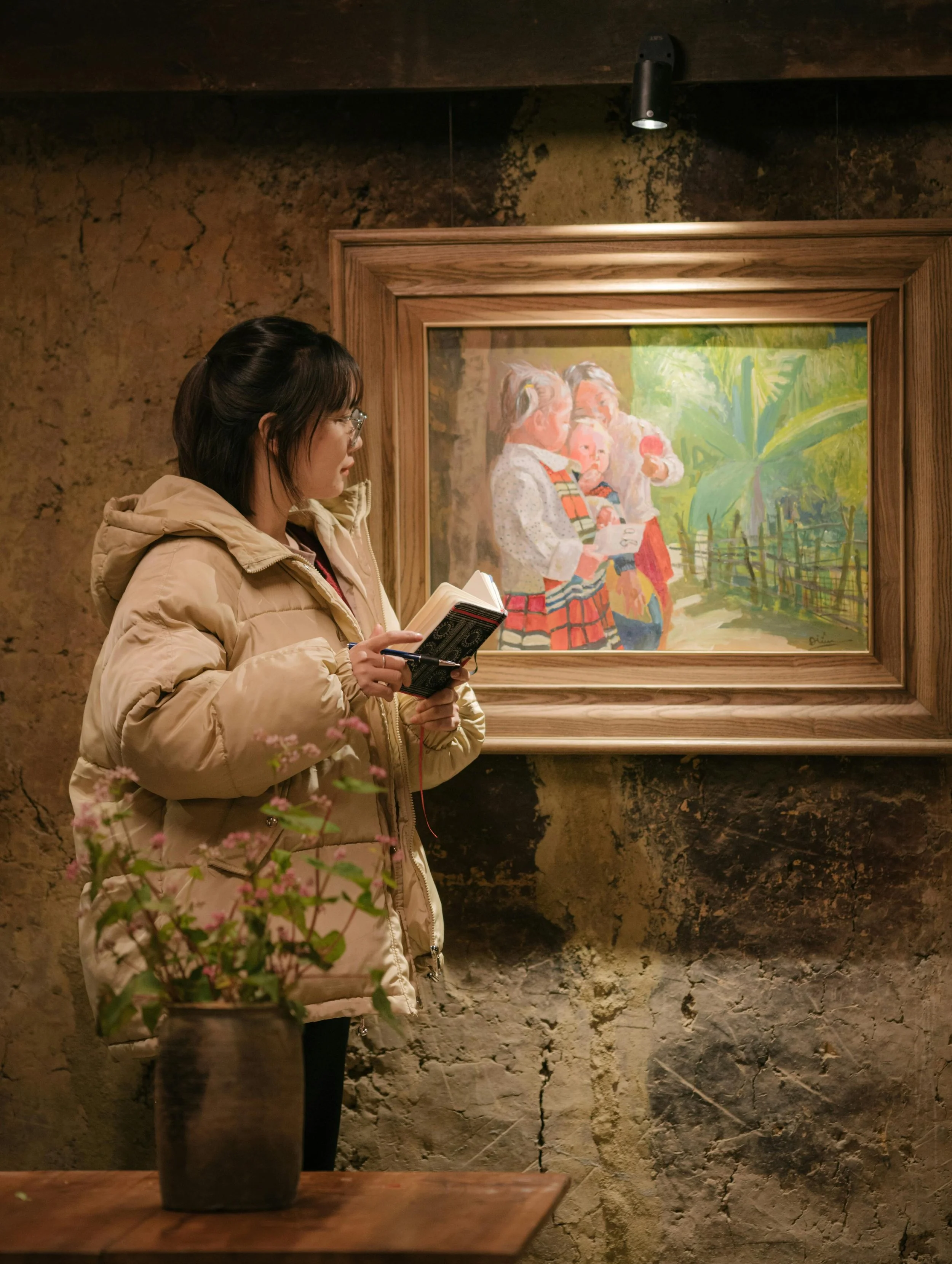 A woman in a beige puffer jacket standing in an art gallery, looking at a framed painting of children outdoors. She holds a small notebook and pen, with a potted plant on a wooden table nearby.