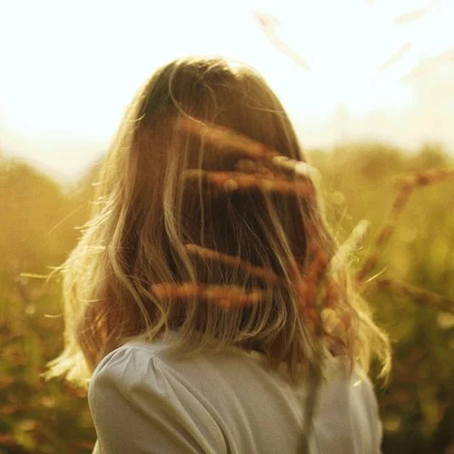 Una ragazza con i capelli mossi che si copre il volto con la mano di fronte a un paesaggio all'aperto durante il tramonto.