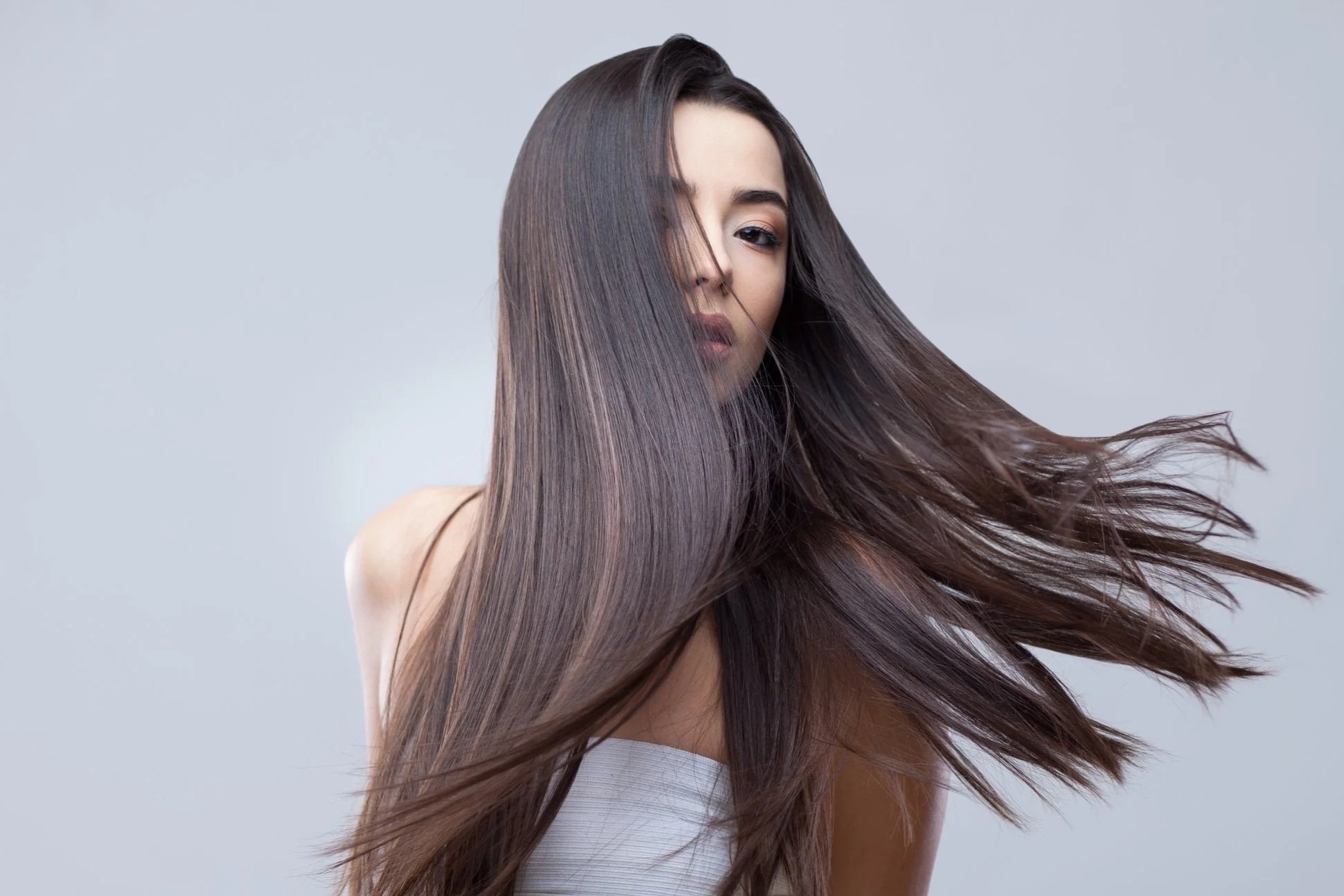 Donna con lunghi capelli castani lisci che si muovono al vento, indossa una maglietta bianca e sfuma in uno sfondo grigio.