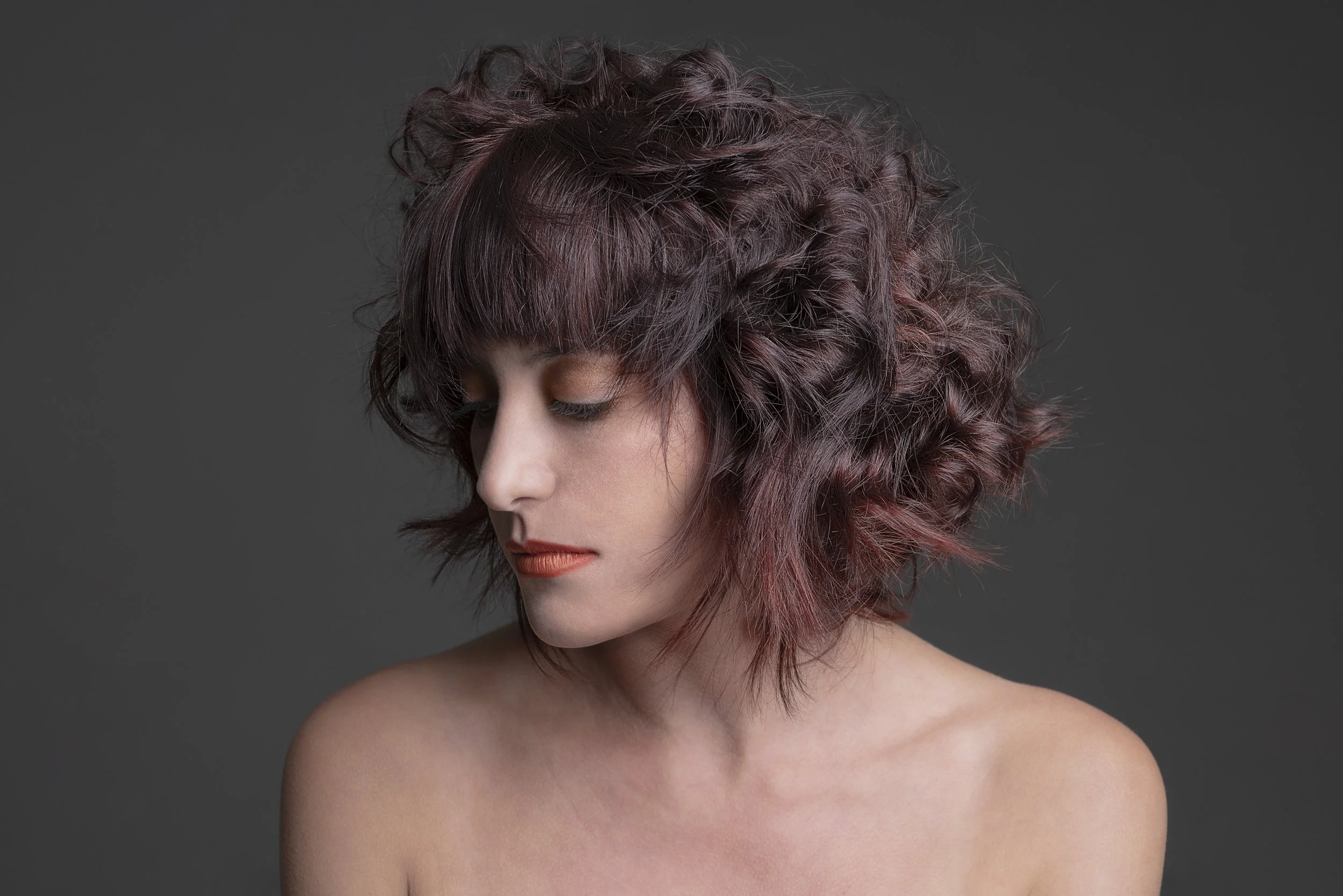 Ragazza con capelli ricci e castani, con trucco naturale e sfondo grigio scuro.