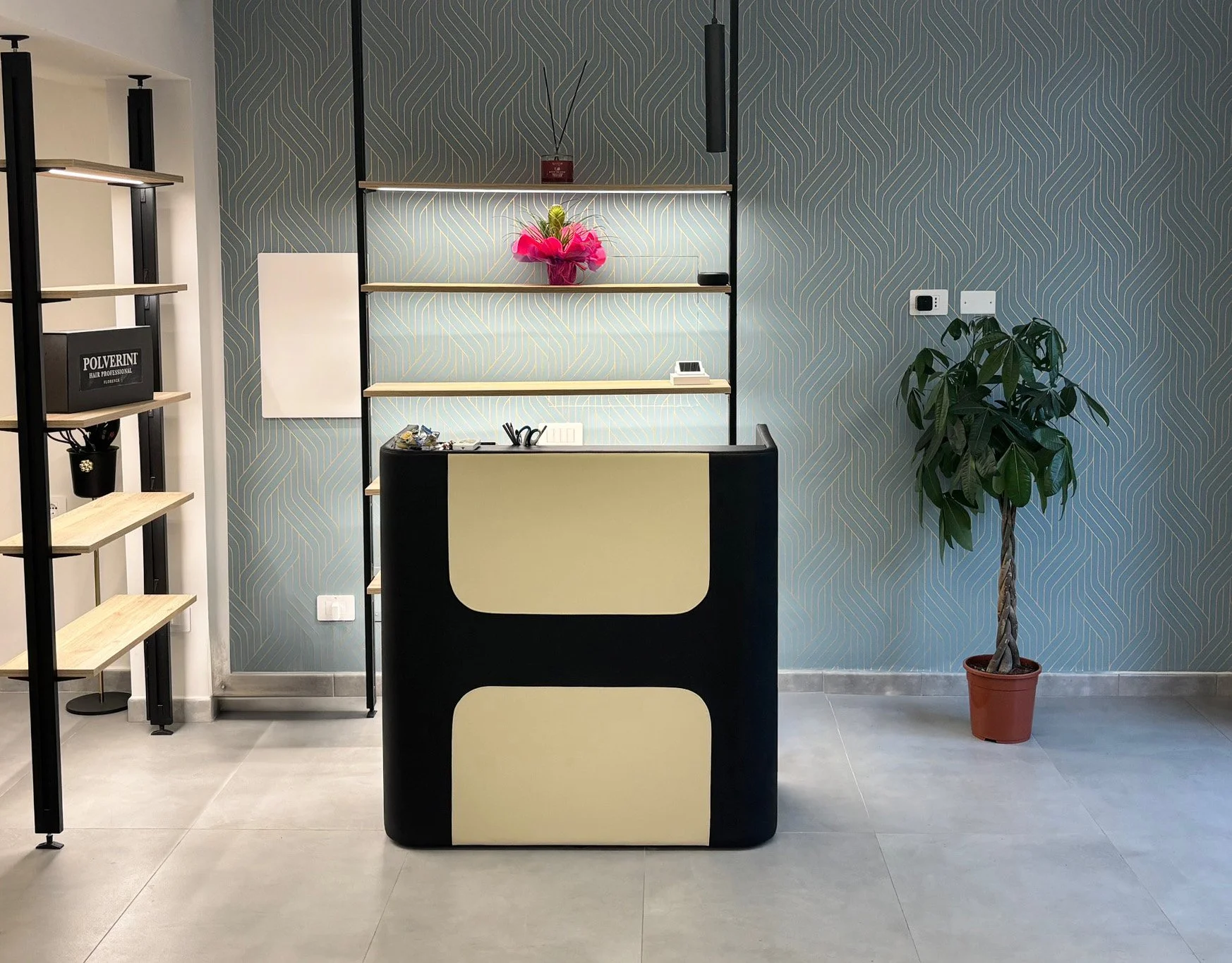 Reception con scrivania nera con dettagli beige, scaffale con oggetti decorativi e piante, pareti con carta da parati a motivi geometrici