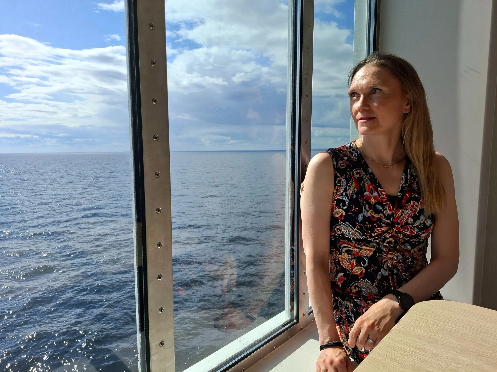 Mari Kalliolahti-Oja istuu ikkunan äärellä, taustalla näkyy suuri meri ja sininen taivas pilvineen.
