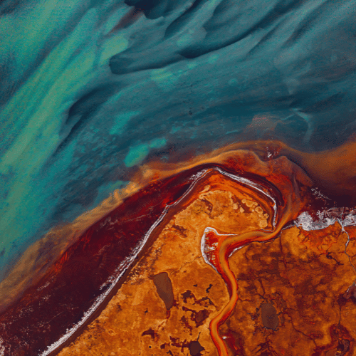 Aerial view of colorful geological formations and water with green, blue, orange, and red hues.
