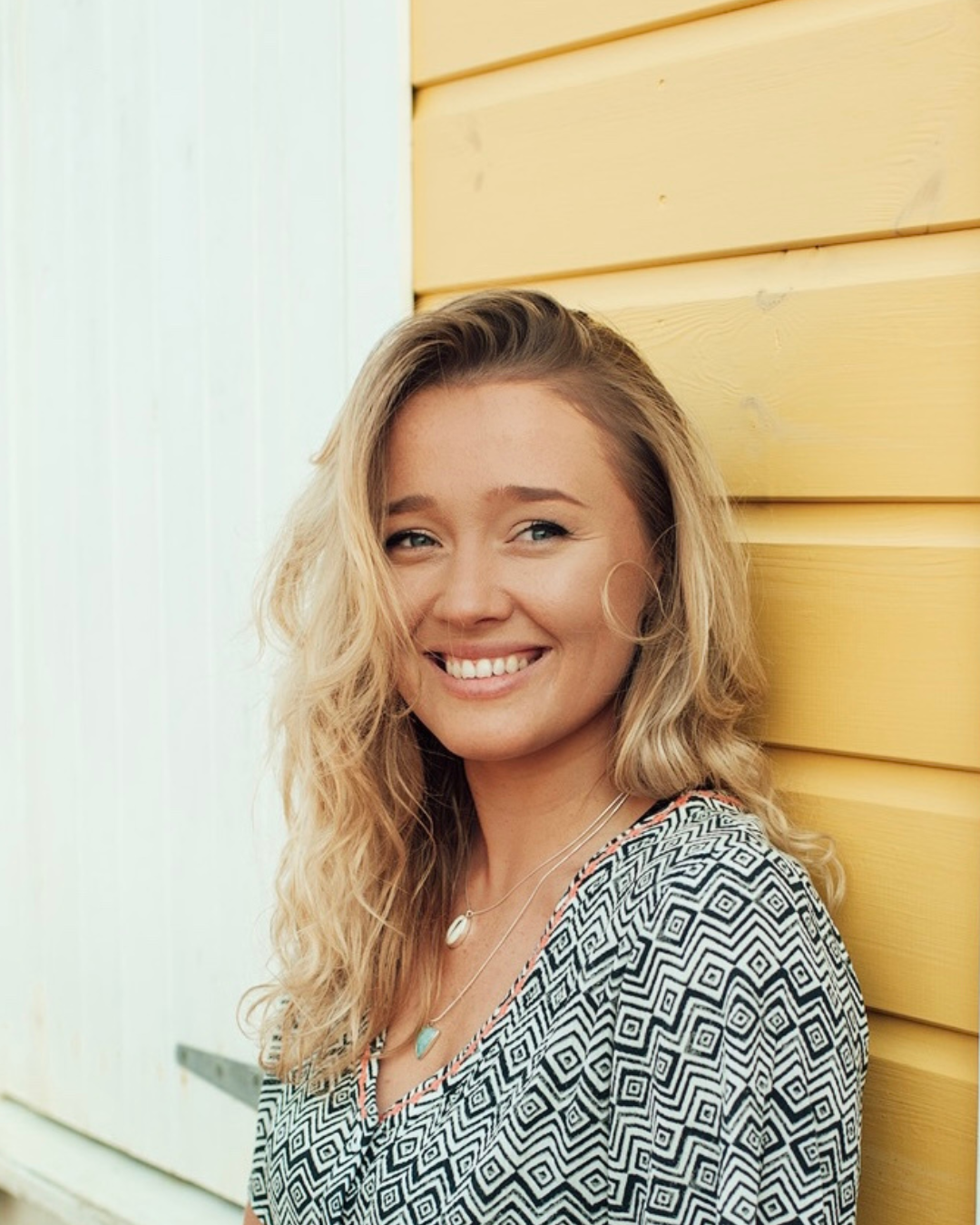 A smiling young woman with wavy blonde hair wearing layered necklaces and a patterned top, leaning against a yellow wooden wall outside.