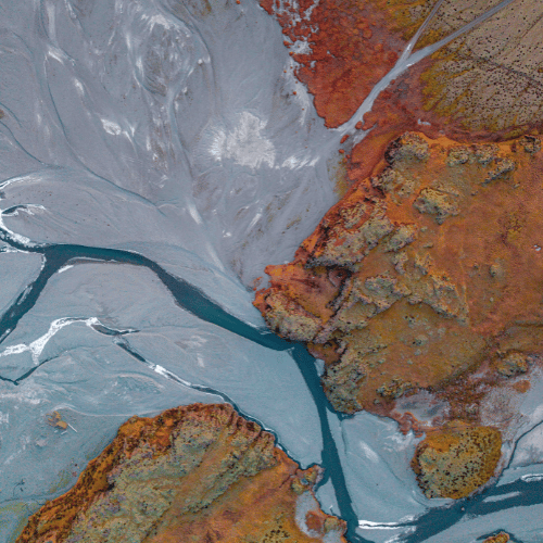 Aerial view of a landscape with a dry riverbed with exposed rocks and sediment in shades of orange, brown, and gray, and patches of pale blue and gray soil.
