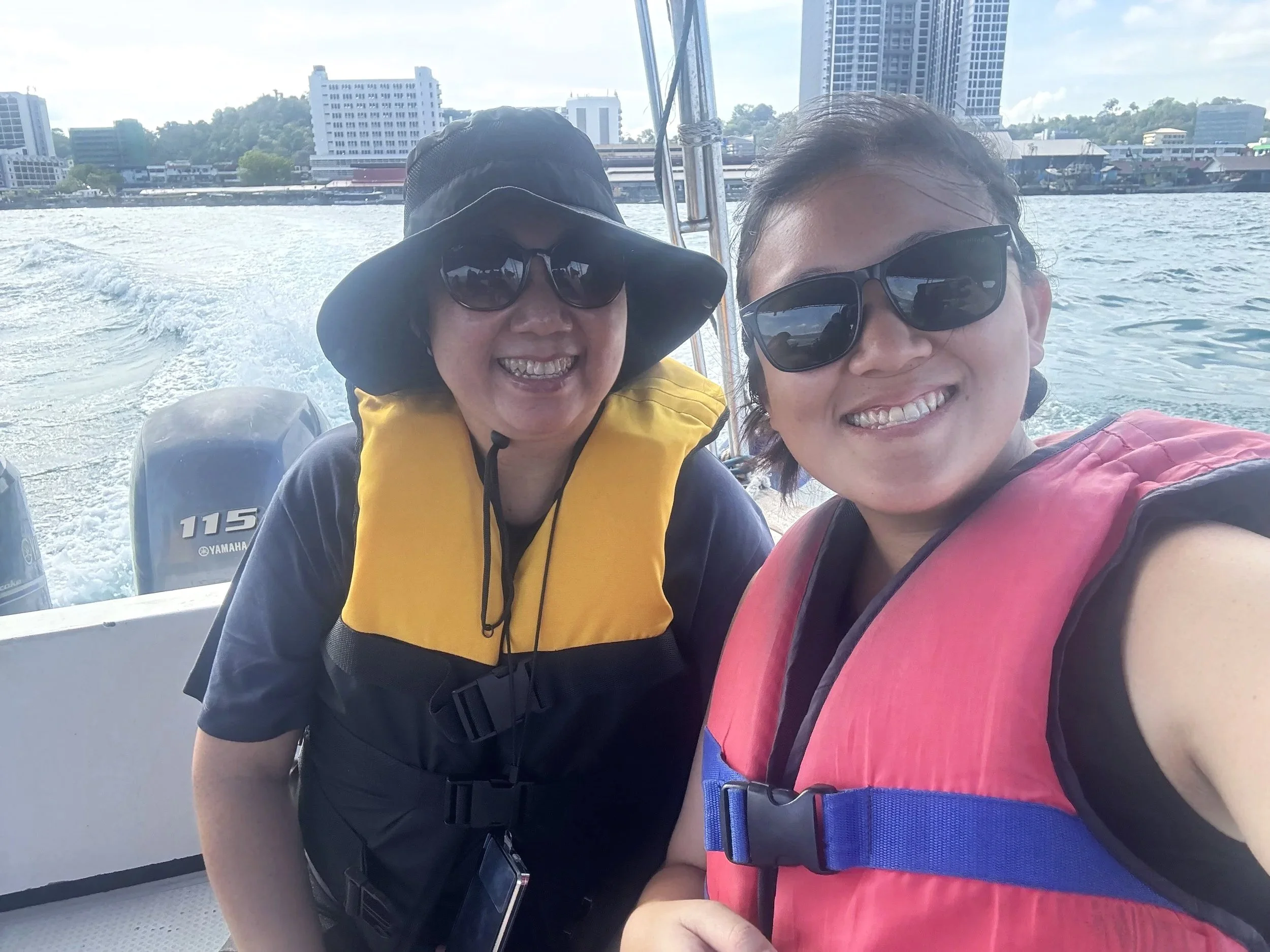 Two women on a boat wearing life jackets and sunglasses, smiling, with city buildings and water in the background.