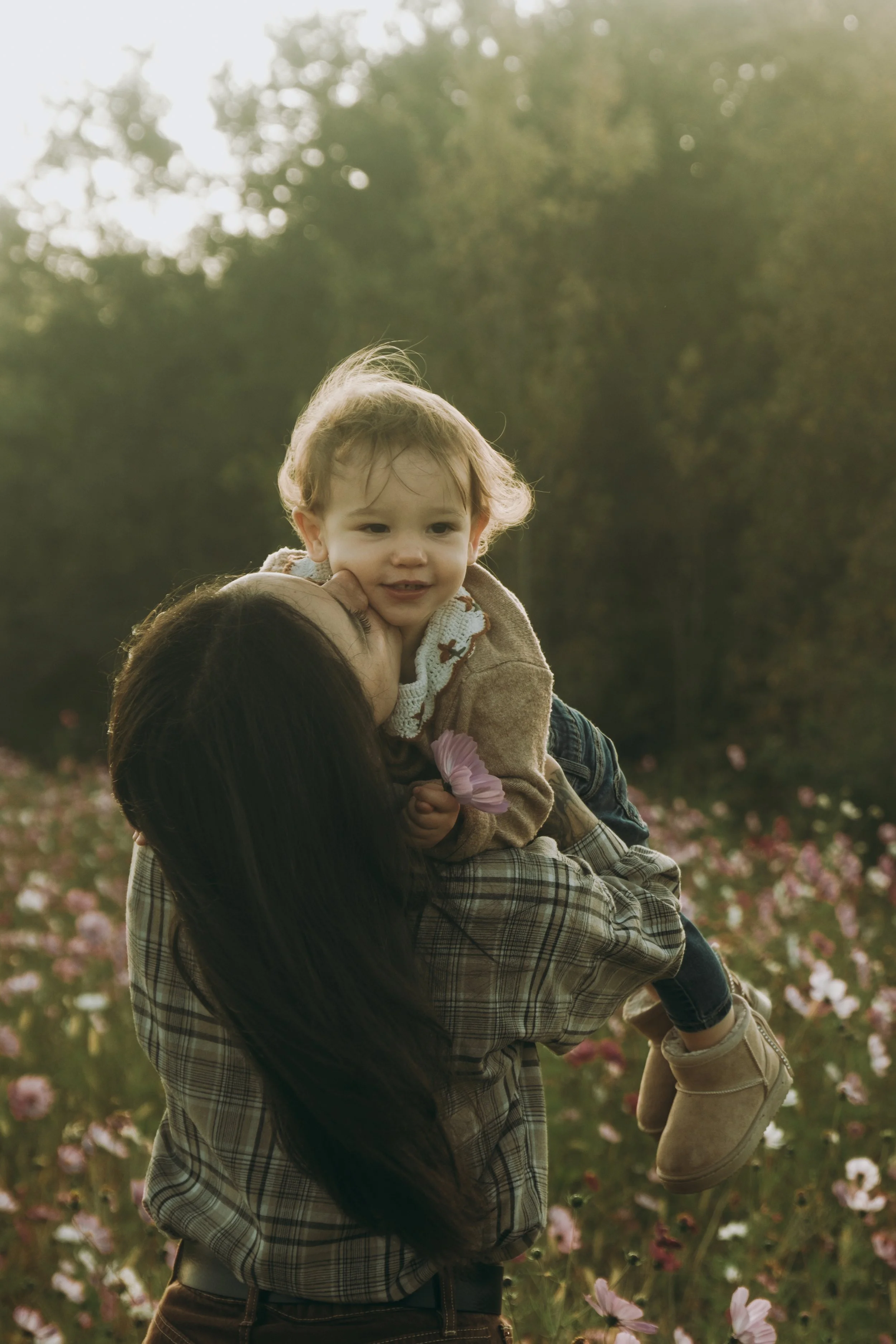 Une femme tient un jeune enfant sur ses épaules dans un champ de fleurs, en plein air avec des arbres en arrière-plan, au crépuscule ou à la lumière douce du soir.