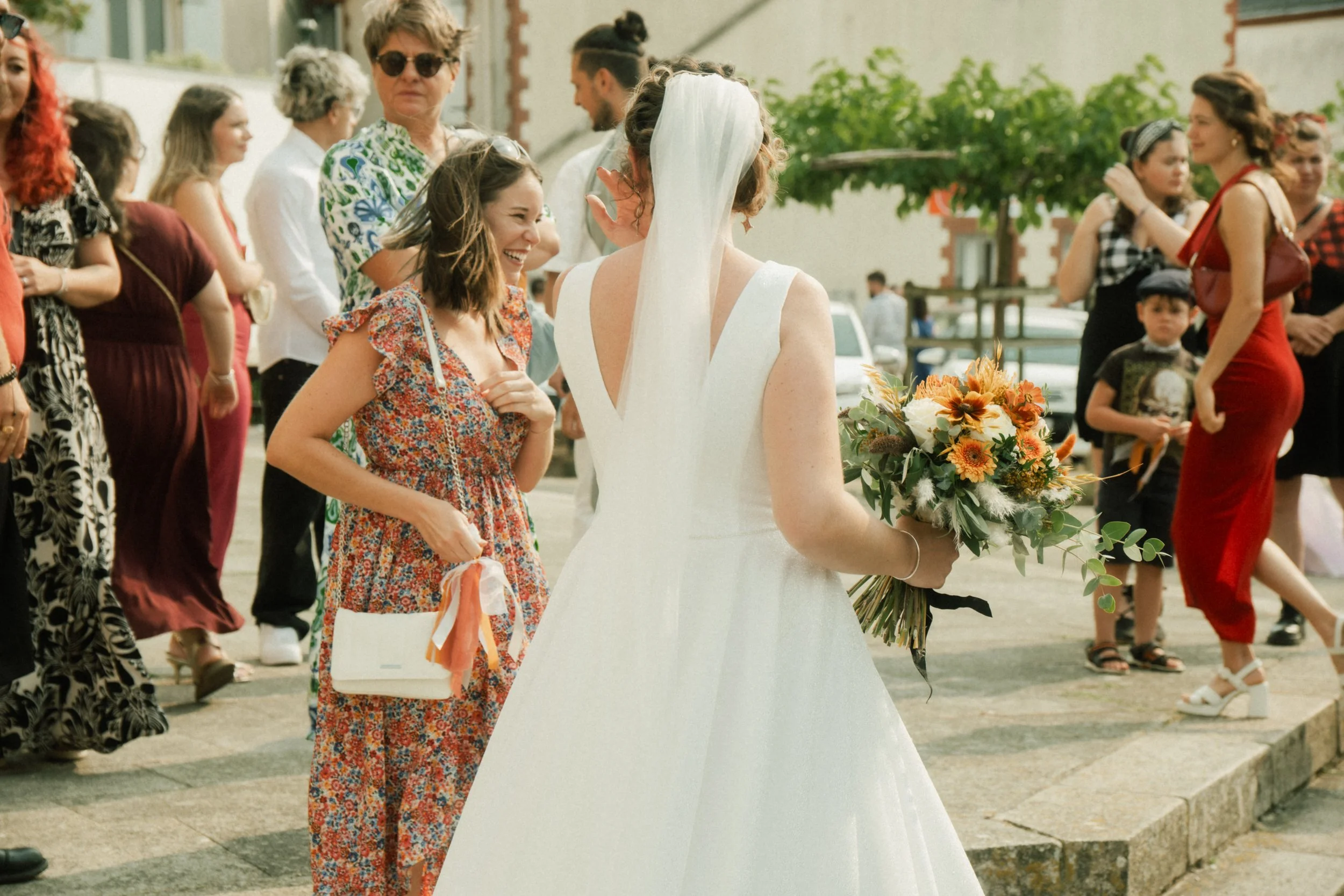 Fête de mariage avec femmes et enfants en tenue colorée, mariée en robe blanche tenant un bouquet de fleurs, sourires et conversations en plein air.