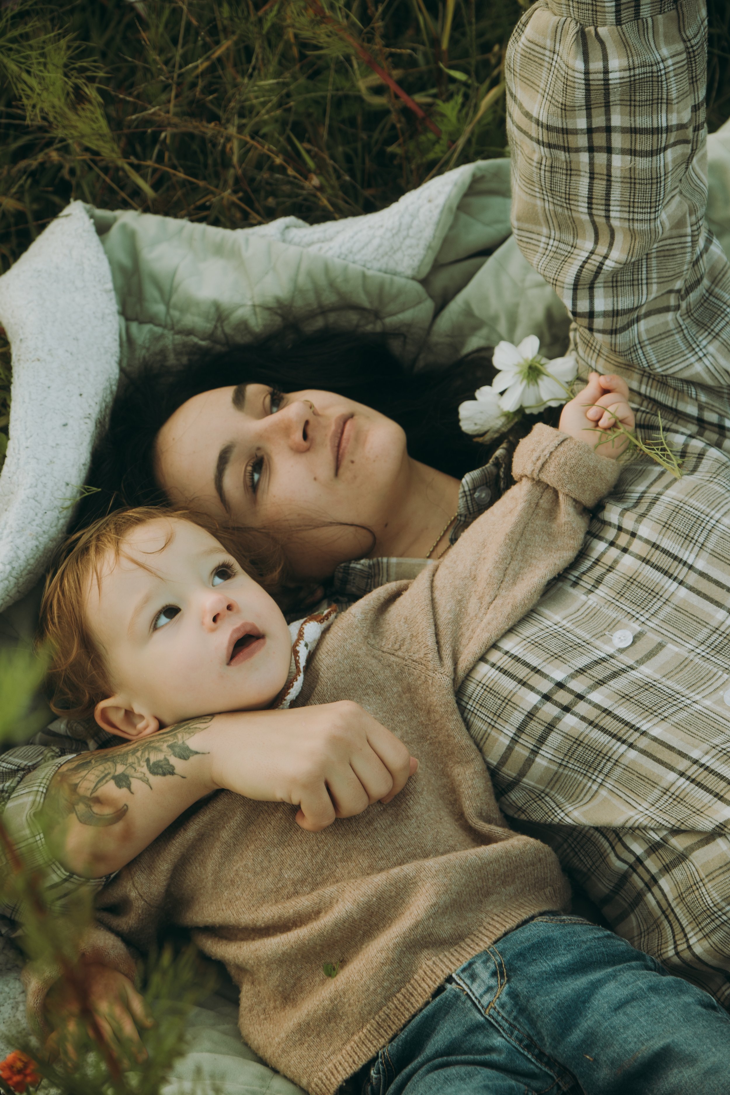 Une femme et un enfant allongés dans l'herbe, la femme tenant une fleur blanche, tous deux regardant vers le haut, entourés de végétation.