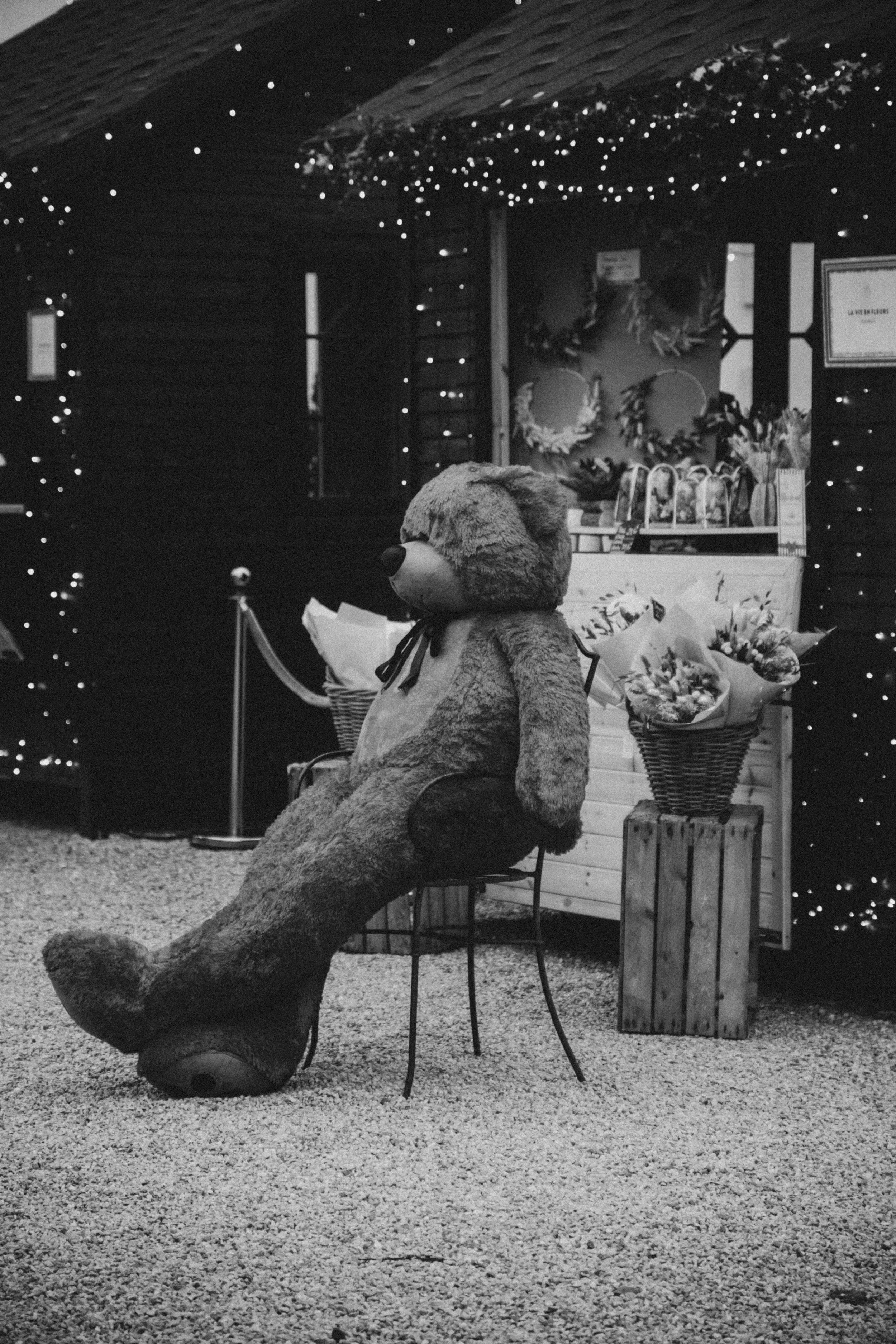 Un grand ours en peluche assis sur une chaise dans un stand décoré avec des guirlandes lumineuses et des bouquets de fleurs, probablement dans un marché ou une fête.