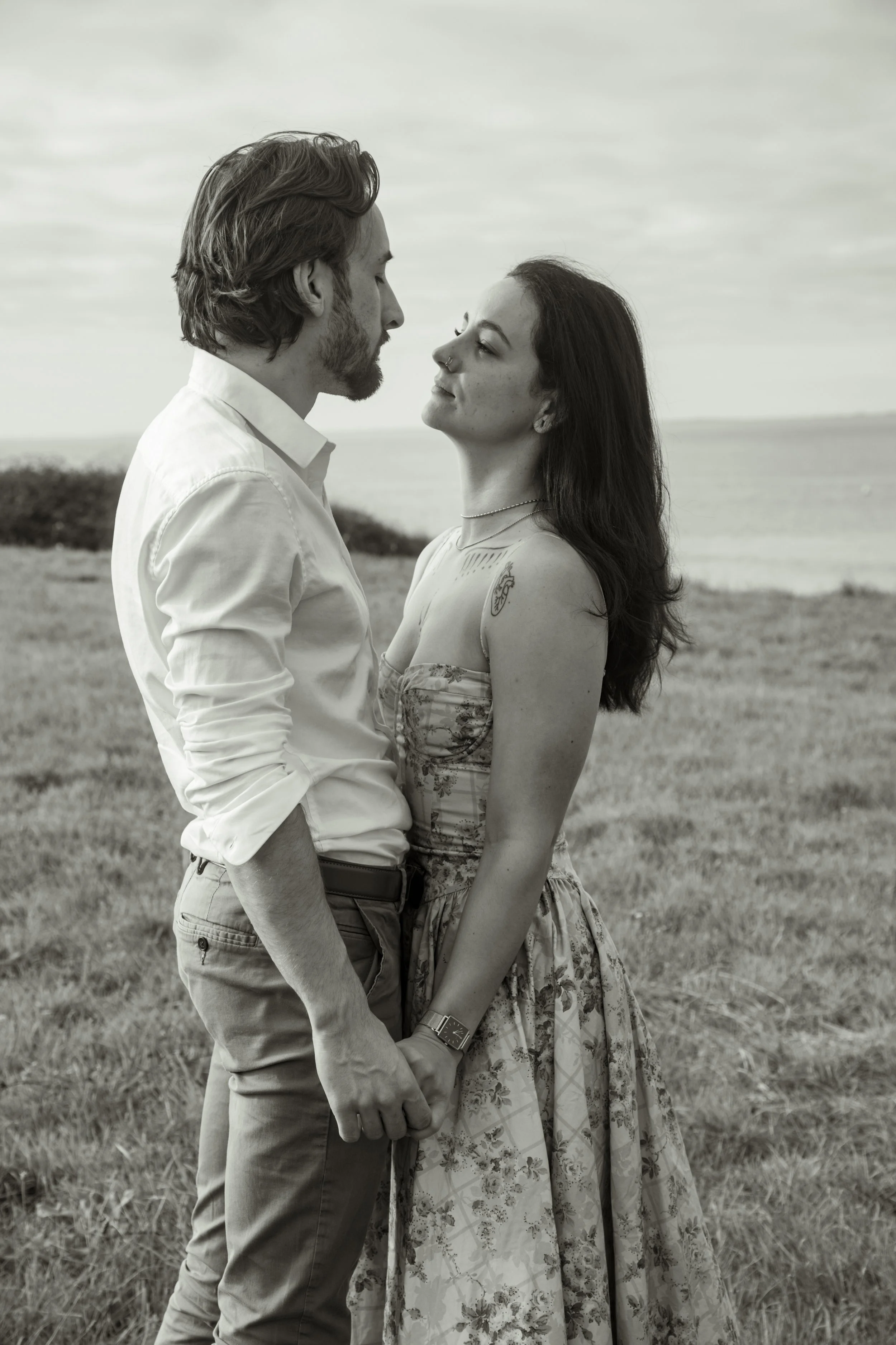 Un couple se tient face à face dans un champ avec la mer en arrière-plan, ils se regardent intensément.