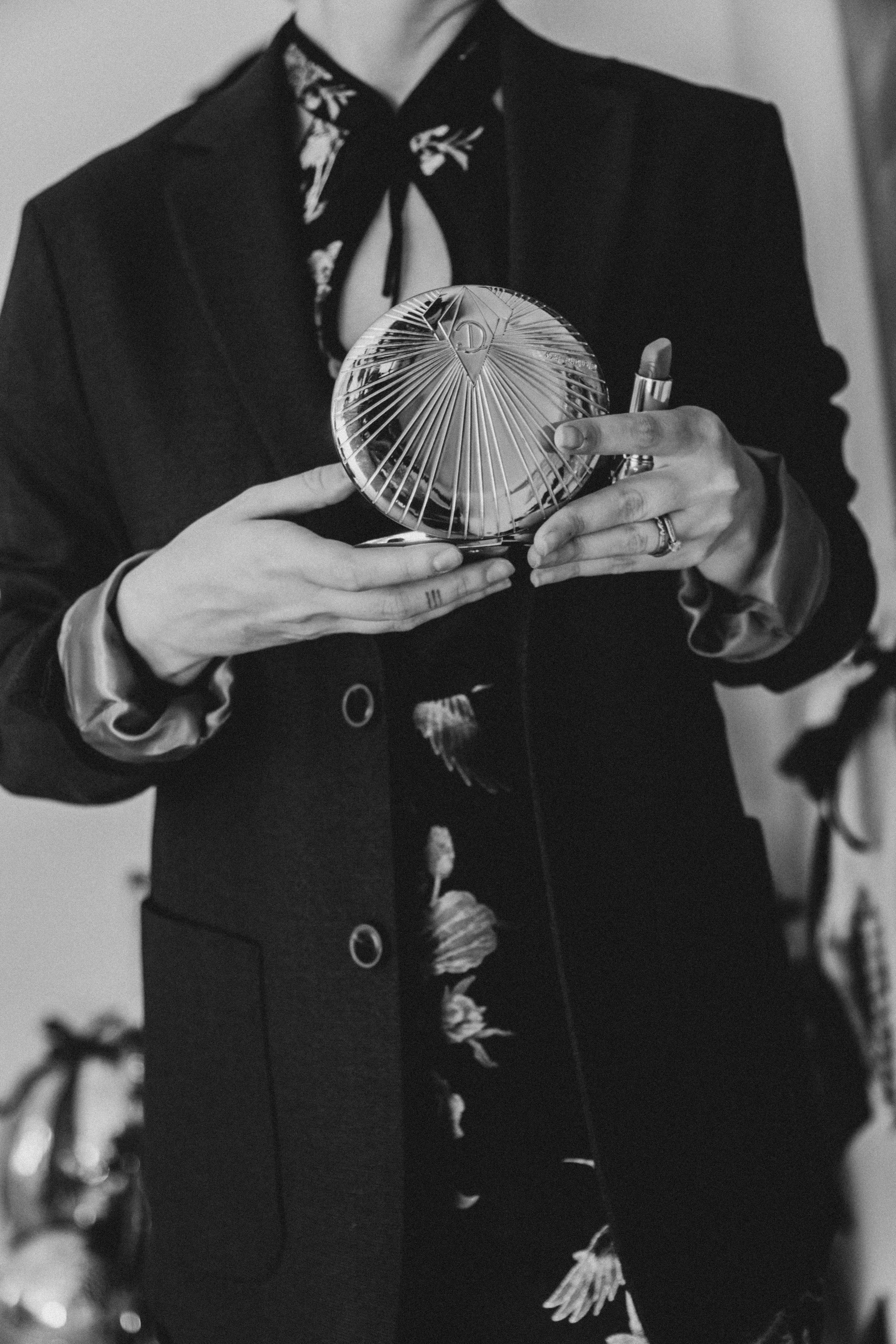 Personne tenant un miroir rond en verre ou en métal, portant un costume noir avec une chemise fleurie et un foulard noir, en noir et blanc.