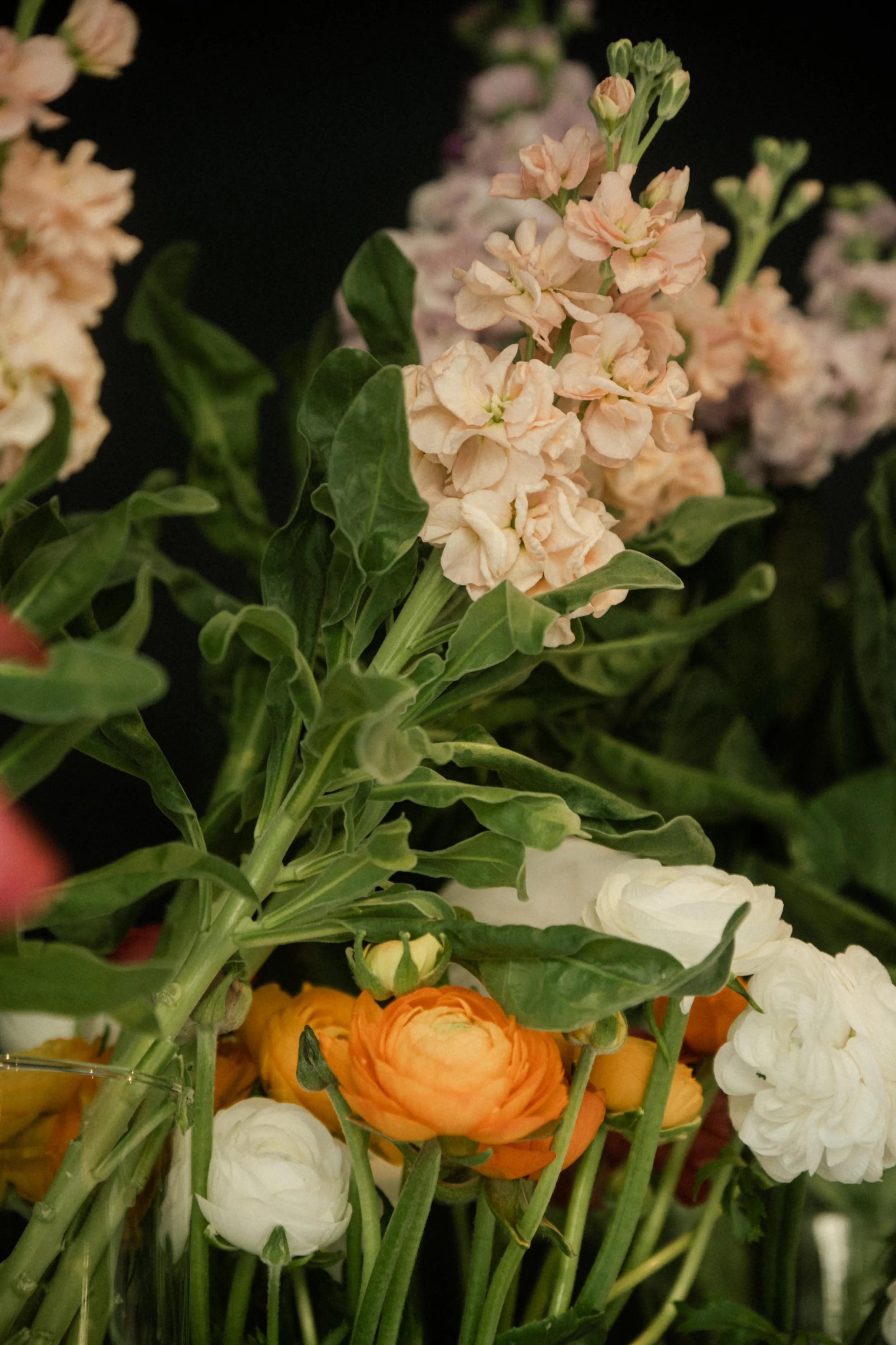Un bouquet de fleurs avec des heuchères en haut, des ranunculus orange et blancs en bas, et des feuillages verts.