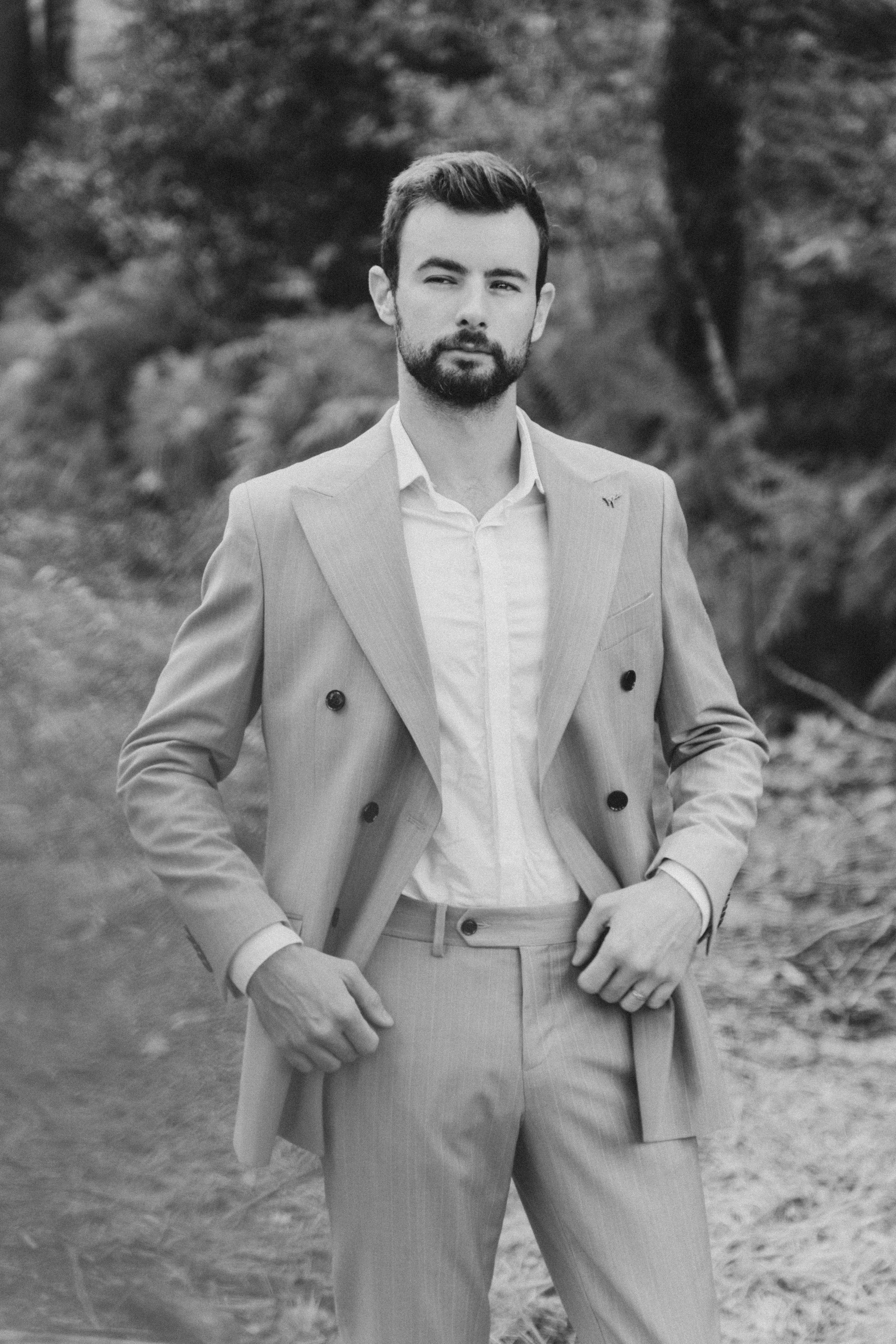 Un homme en costume sans cravate, debout dans la nature, en noir et blanc.