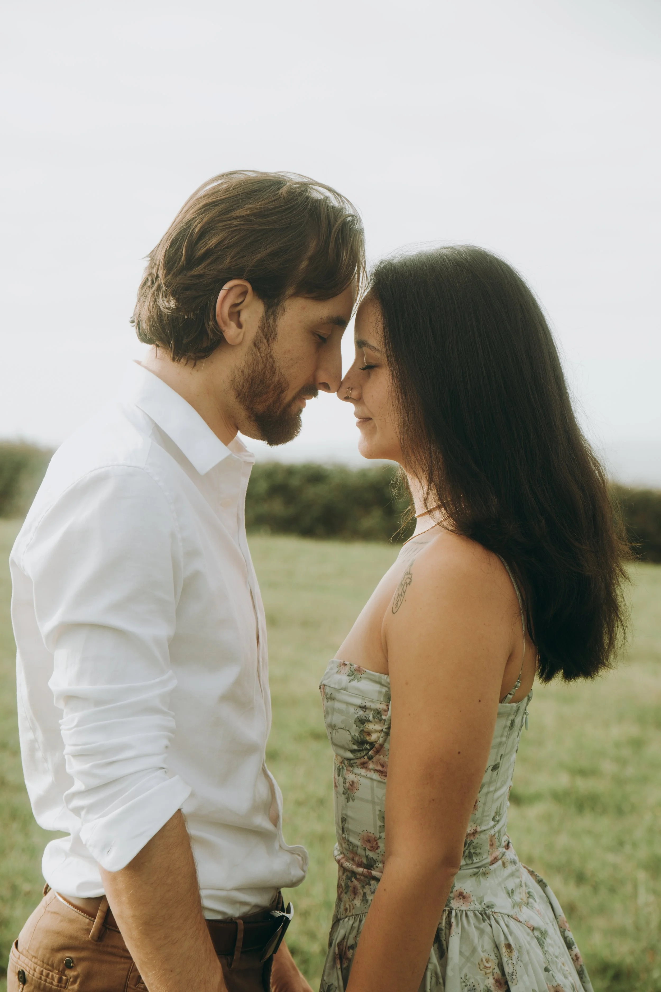 Un couple jeune, hommes et femmes, se tenant face à face avec le front presque touchant dans un champ vert, expression de tendresse, moment intime, nature en arrière-plan.